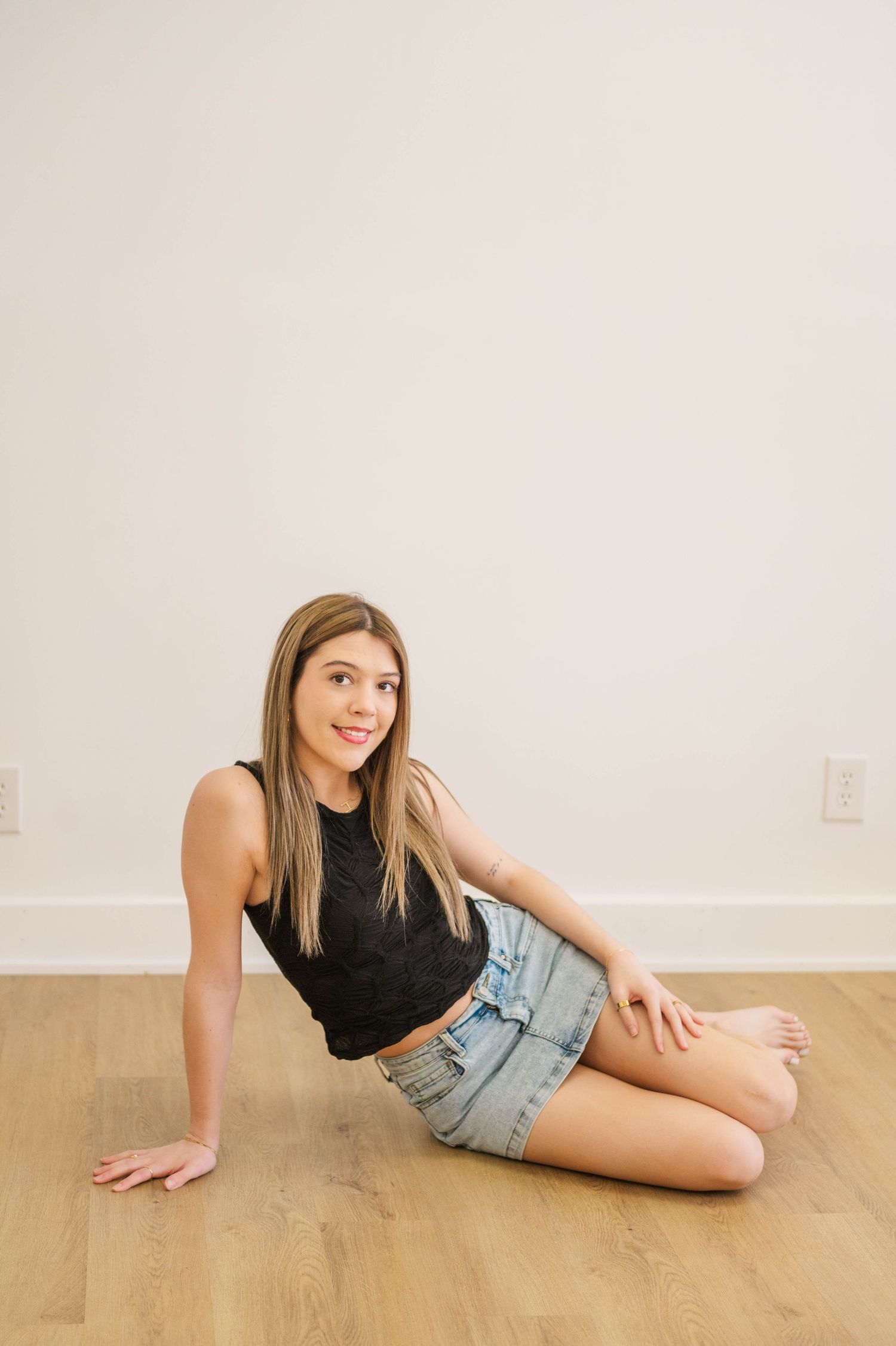Young woman in black sleeveless top and denim mini skirt sitting on hardwood floor against white wall.