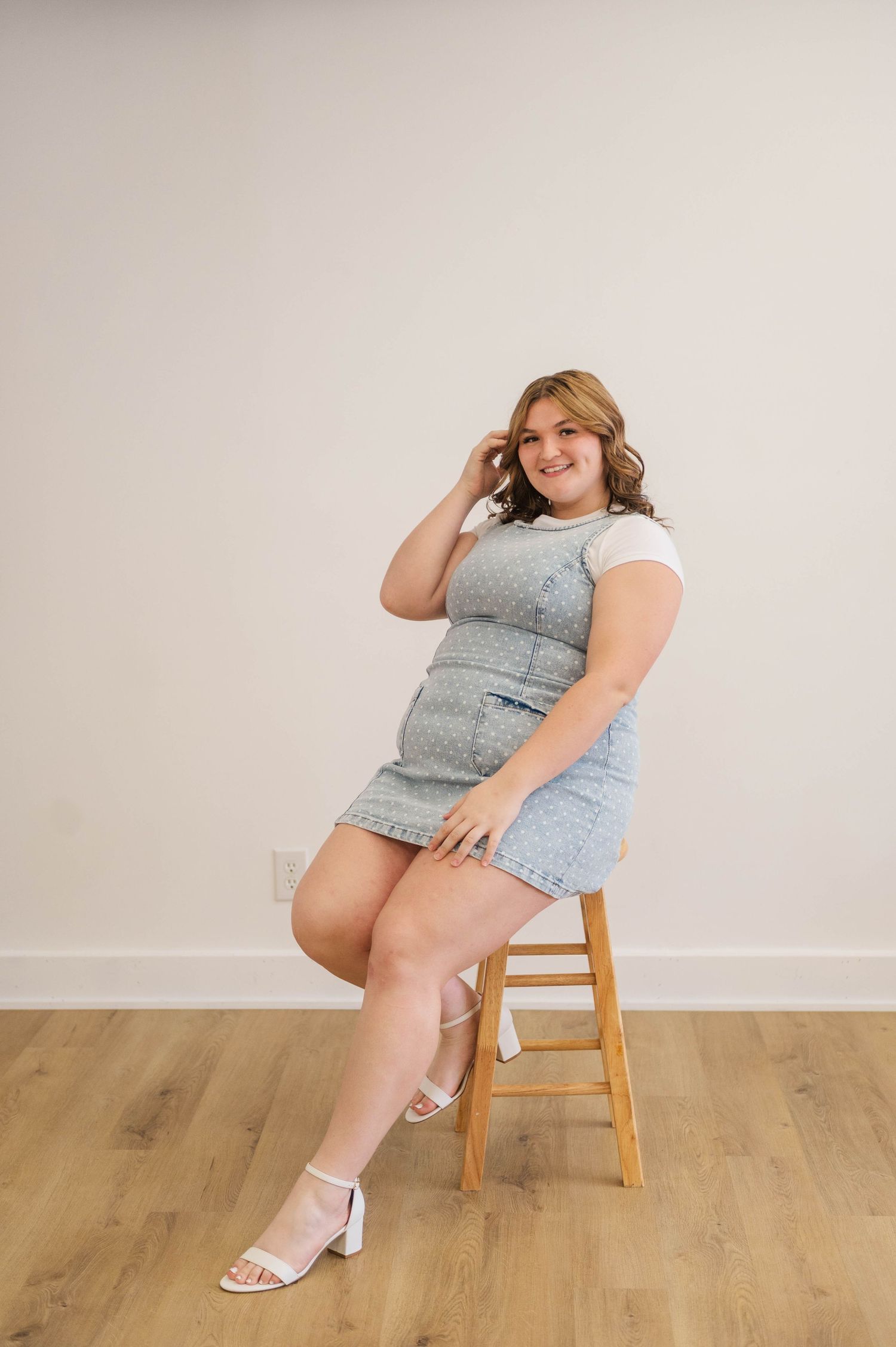 Smiling woman in denim pinafore dress and white heels sitting on wooden stool against white wall.