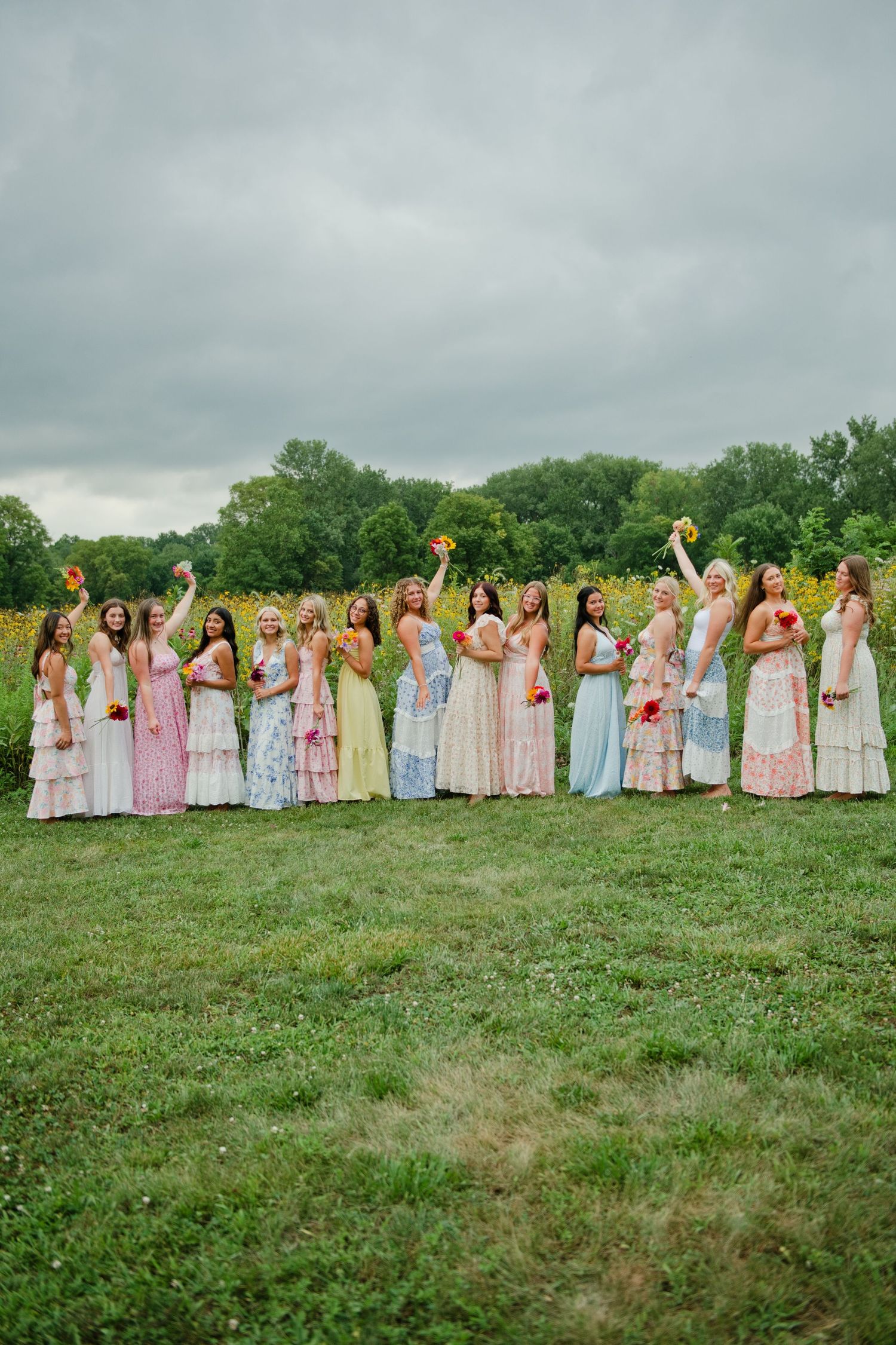 A group of bridesmaids in colorful pastel dresses celebrate in a green field with wildflowers under cloudy skies.