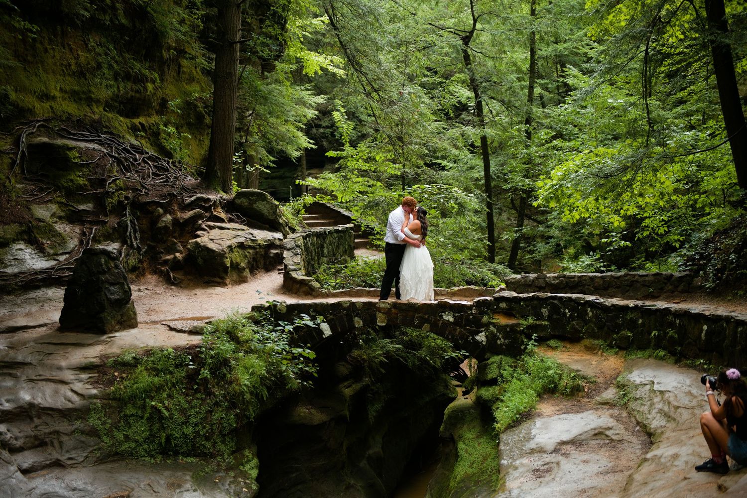 Bride and groom embrace on mossy stone bridge surrounded by lush green forest in a romantic outdoor wedding photo.