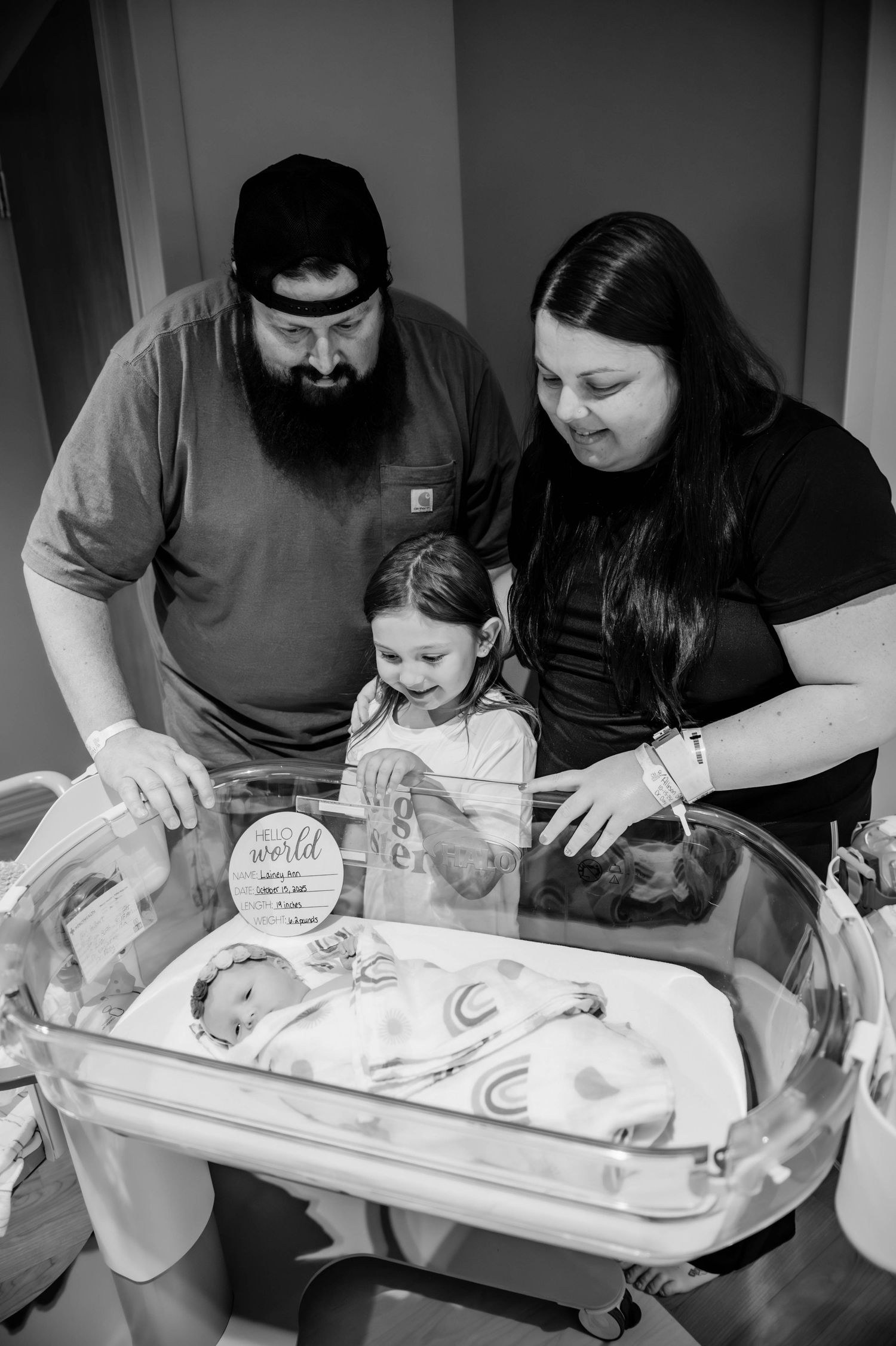 A family gathers around a hospital bassinet to look at a newborn baby in black and white.
