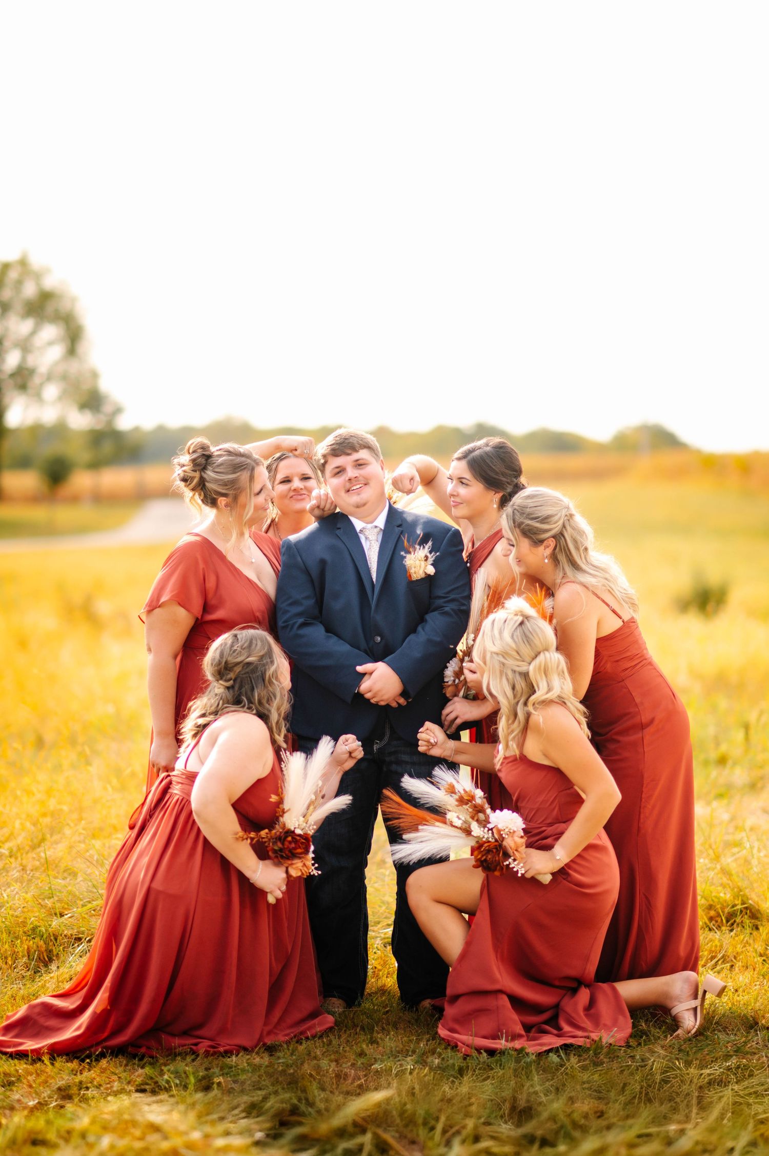 Bridal party in red dresses gather around couple in grassy field at golden hour.