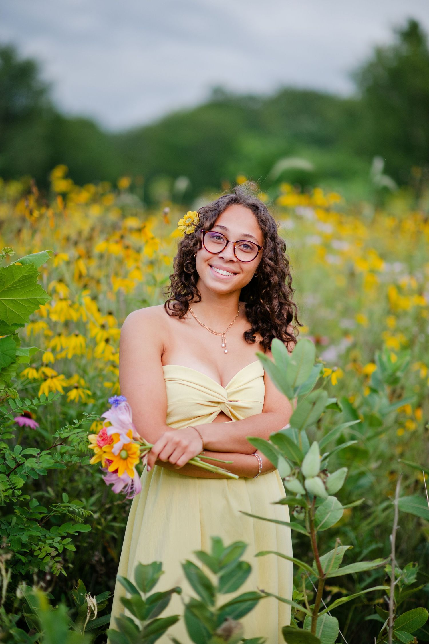 A smiling young woman in a yellow strapless dress holds wildflowers in a vibrant yellow flower field.