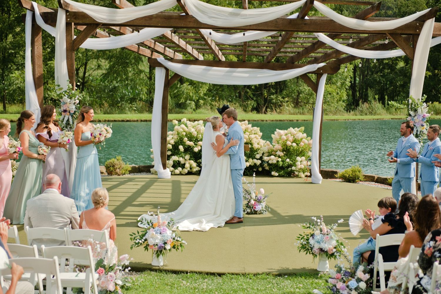 Couple shares first kiss at outdoor wedding ceremony under floral pergola beside serene pond with guests seated on white chairs.