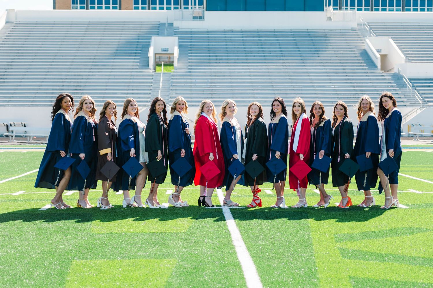 Group of graduates in navy and red gowns posing together on a green football field.