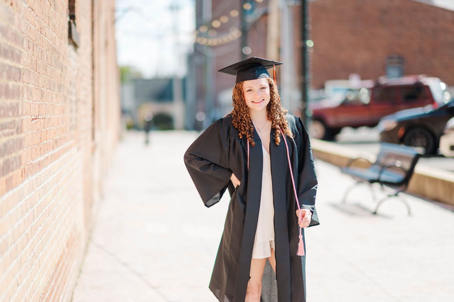 Smiling graduate in black cap and gown posing on urban brick sidewalk.