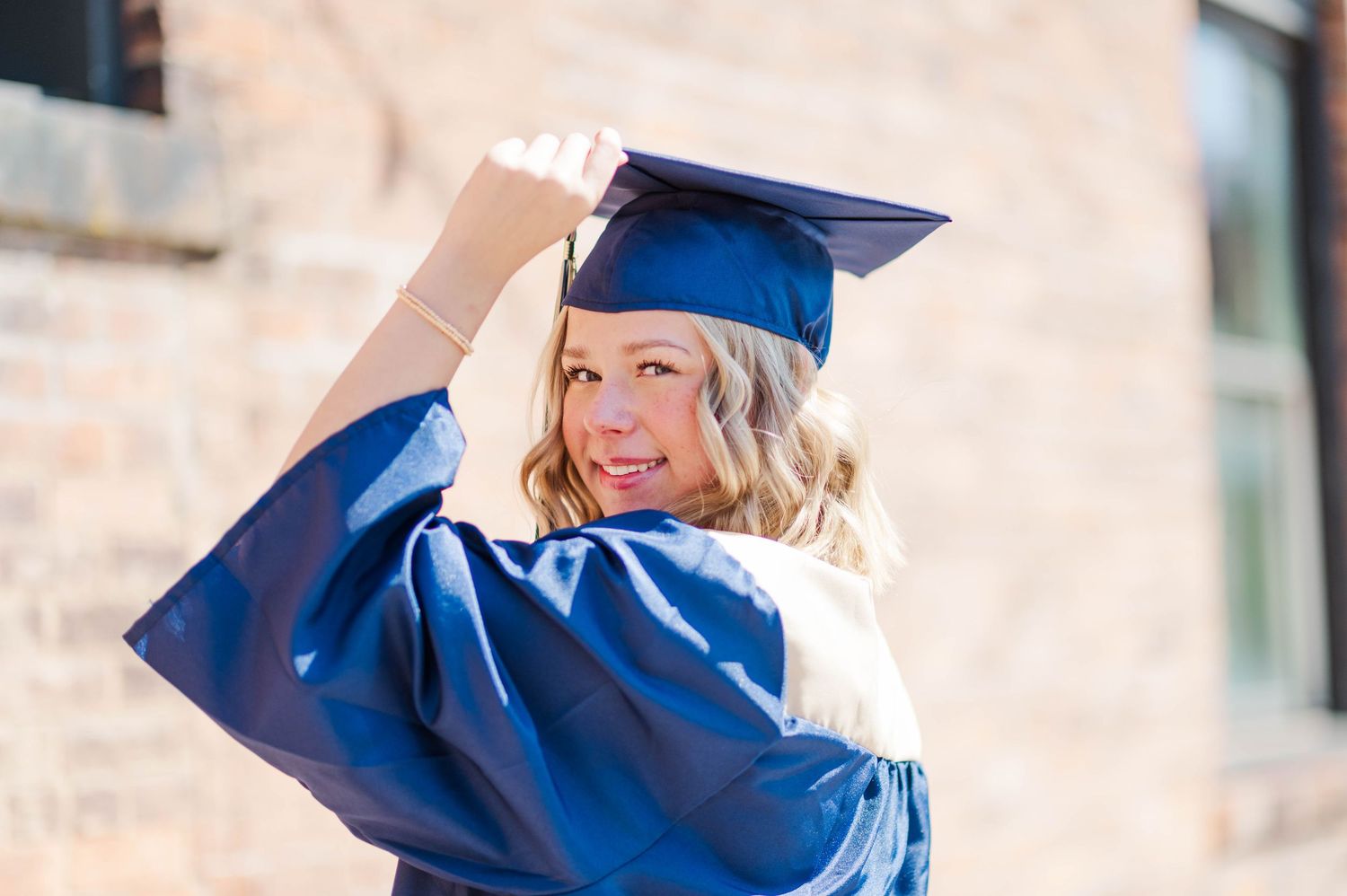 Young woman in blue graduation cap and gown posing outdoors near a brick building.