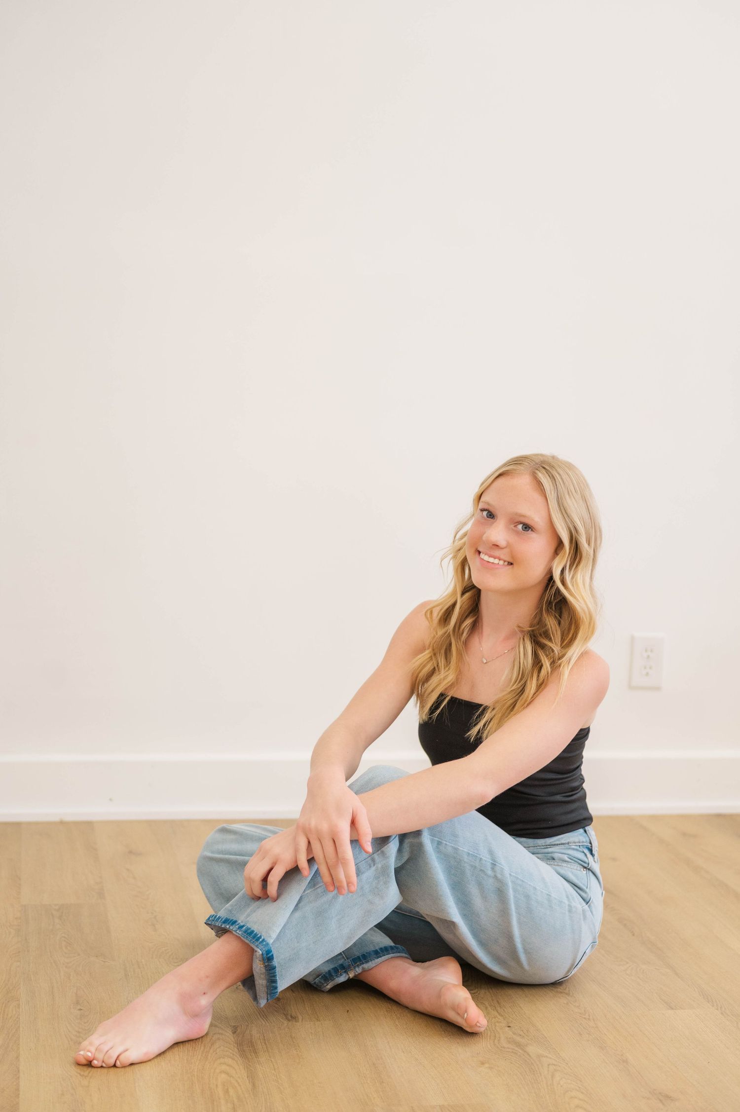 Blonde woman sitting barefoot on hardwood floor smiling, wearing black tank top and light blue jeans.