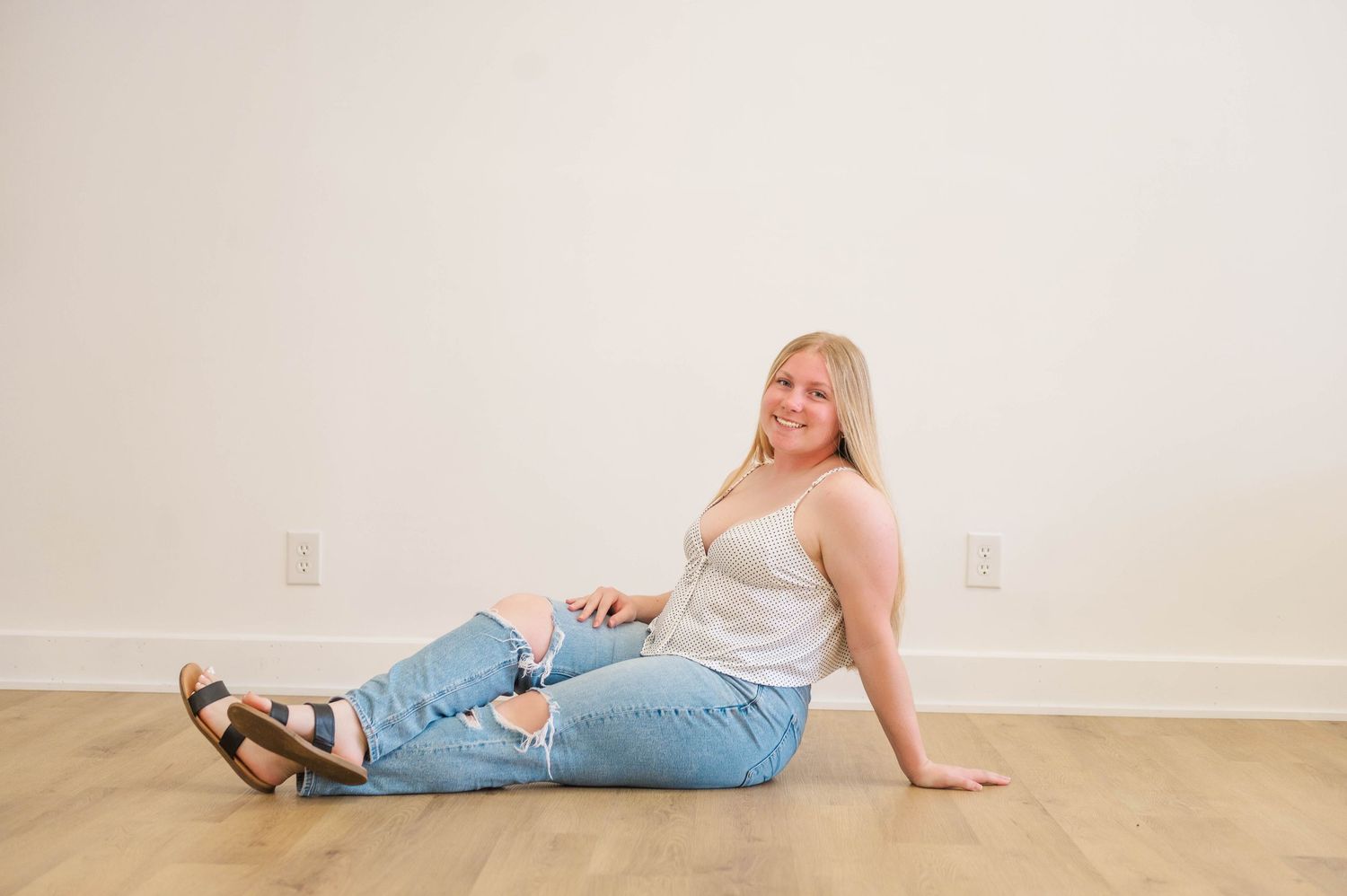 Blonde woman in white tank and ripped jeans sitting on hardwood floor, smiling casually.