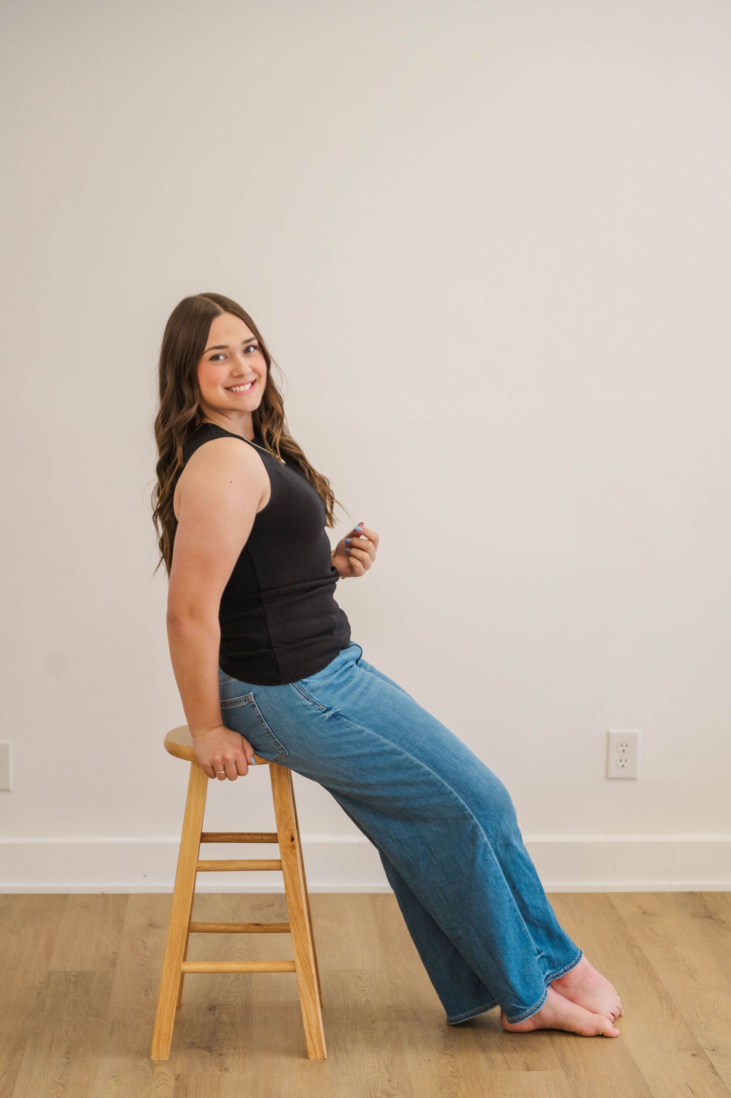 Brunette woman in black tank top and wide-leg jeans sitting sideways on a wooden stool.