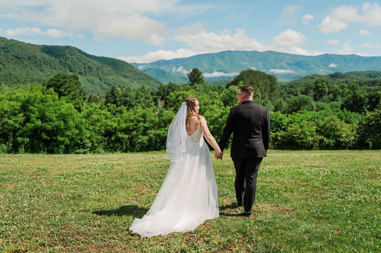 Bride and groom holding hands walking through green meadow with stunning Smoky Mountain views on their wedding day.