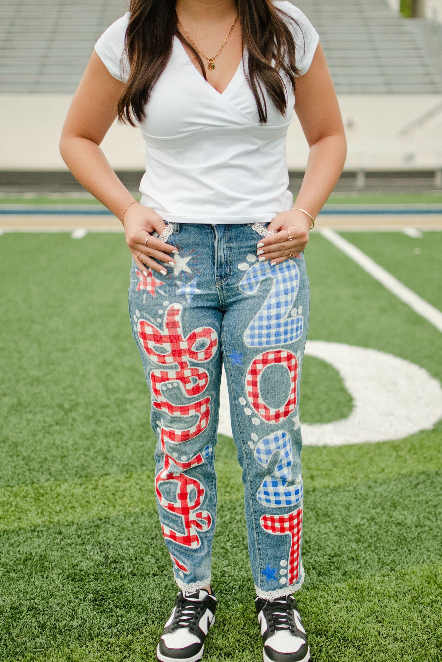 Close-up of teen girl wearing custom painted senior 2025 red gingham patterned jeans on a football field.