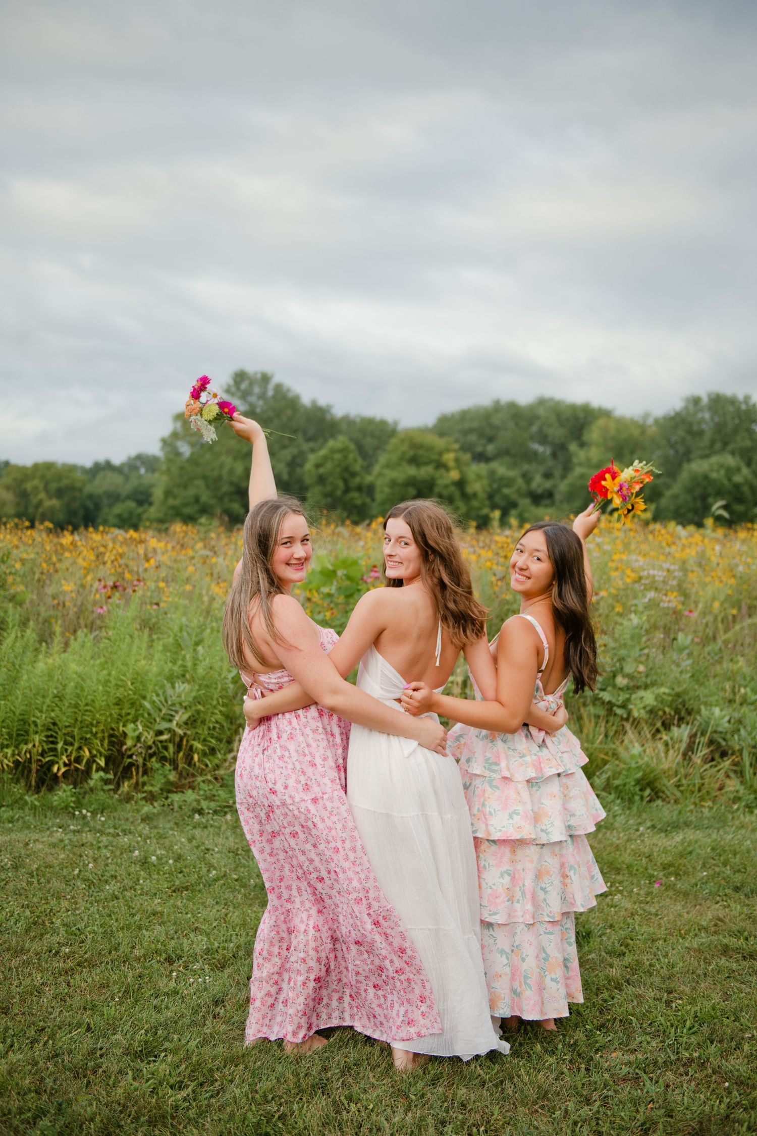 Three bridesmaids in pink and white dresses laughing and raising flower bouquets in a sunny field.