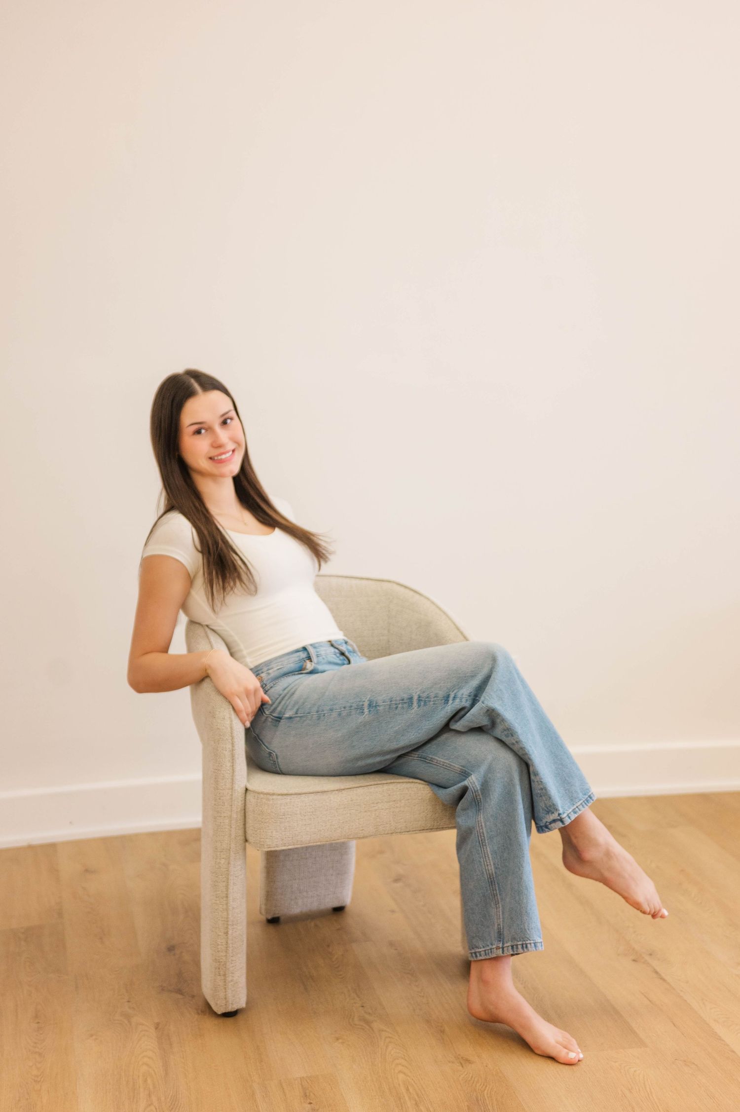 Brunette woman in white tee and blue jeans sitting relaxed barefoot in beige chair on hardwood floor.