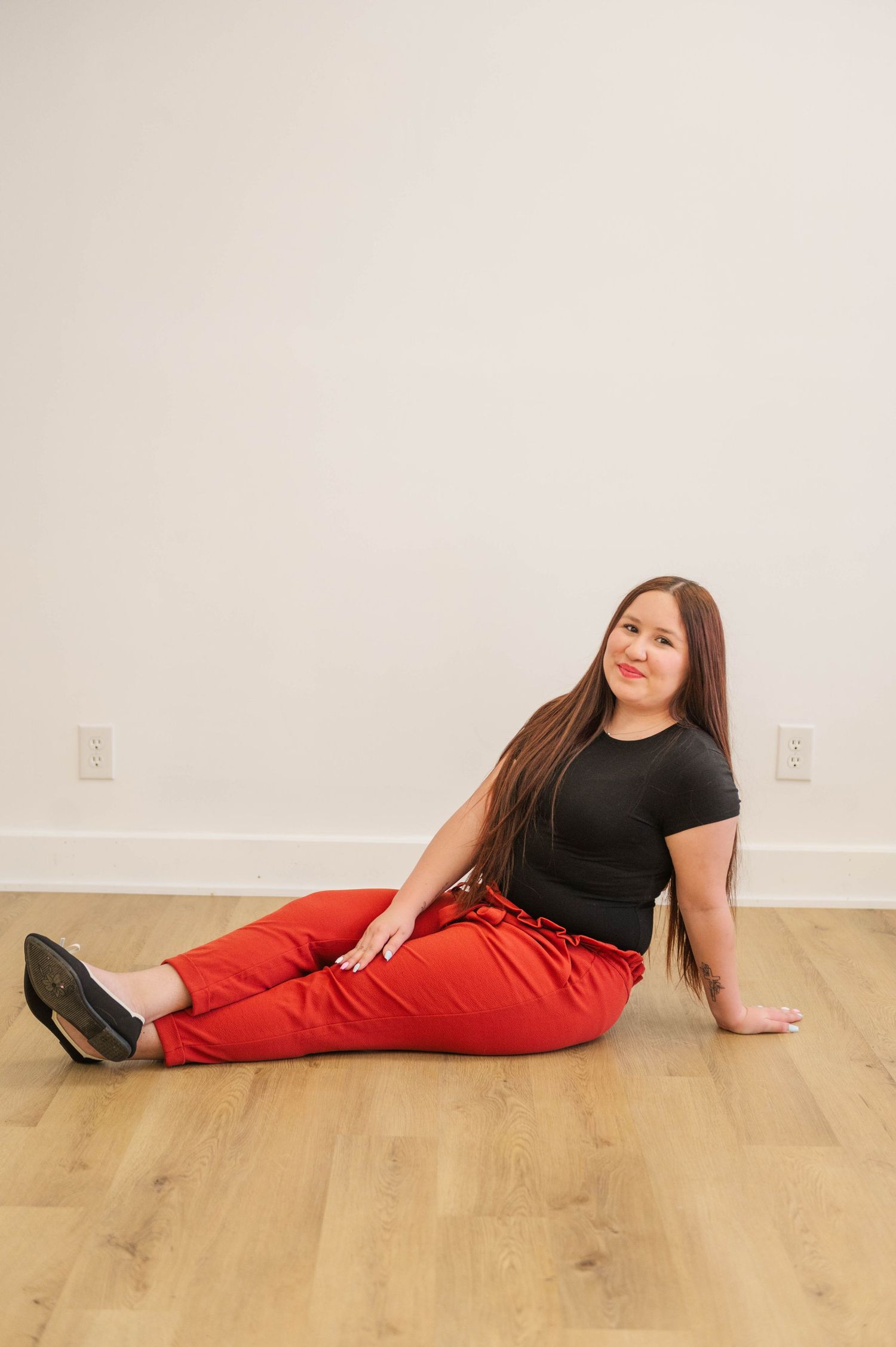 Woman with long hair in black tee and red pants sits casually on hardwood floor against white wall.