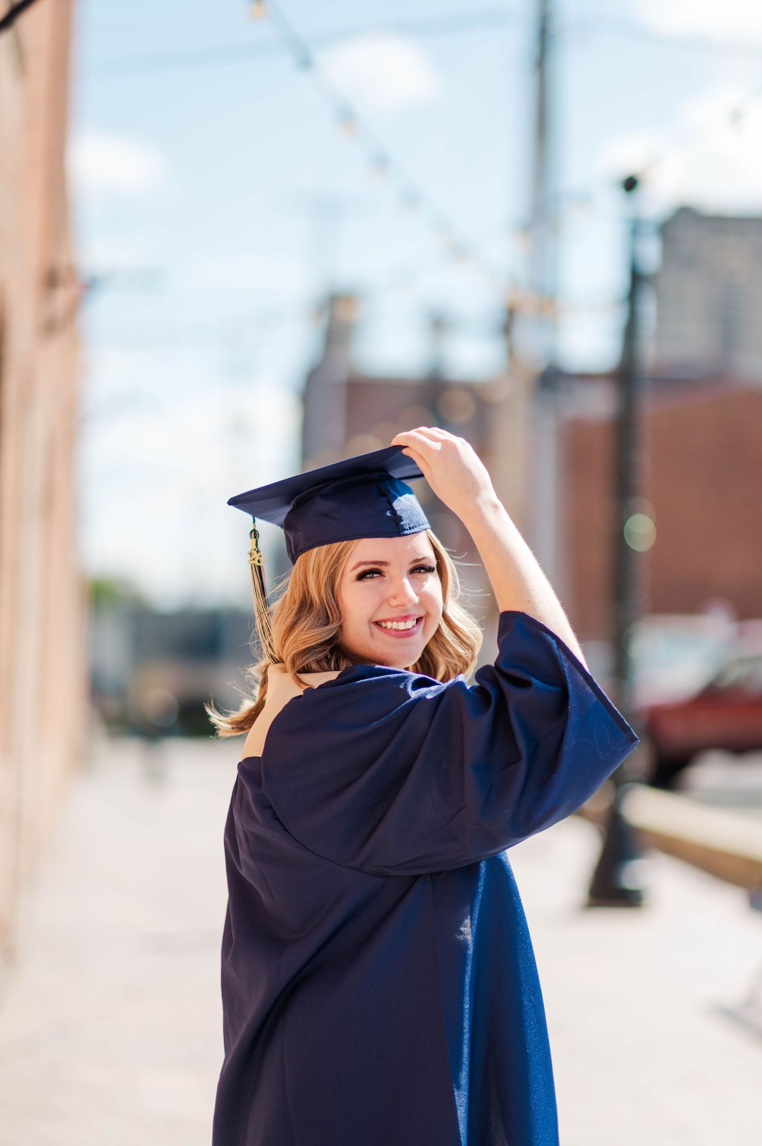 Smiling blonde woman in navy blue graduation cap and gown posing on sunny urban street.