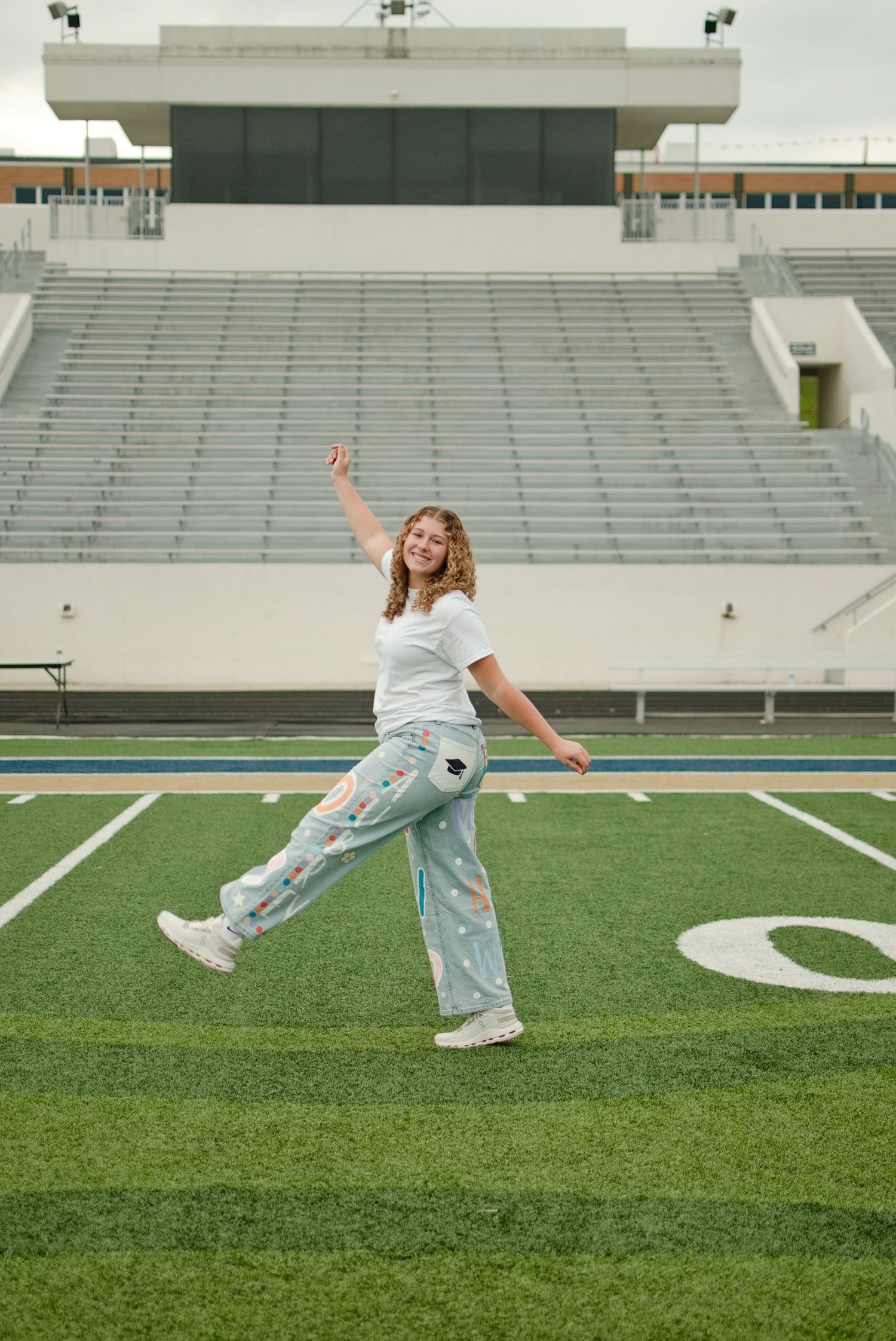 Young woman posing joyfully on football field turf wearing patterned pants and white tee with stadium stands behind her.