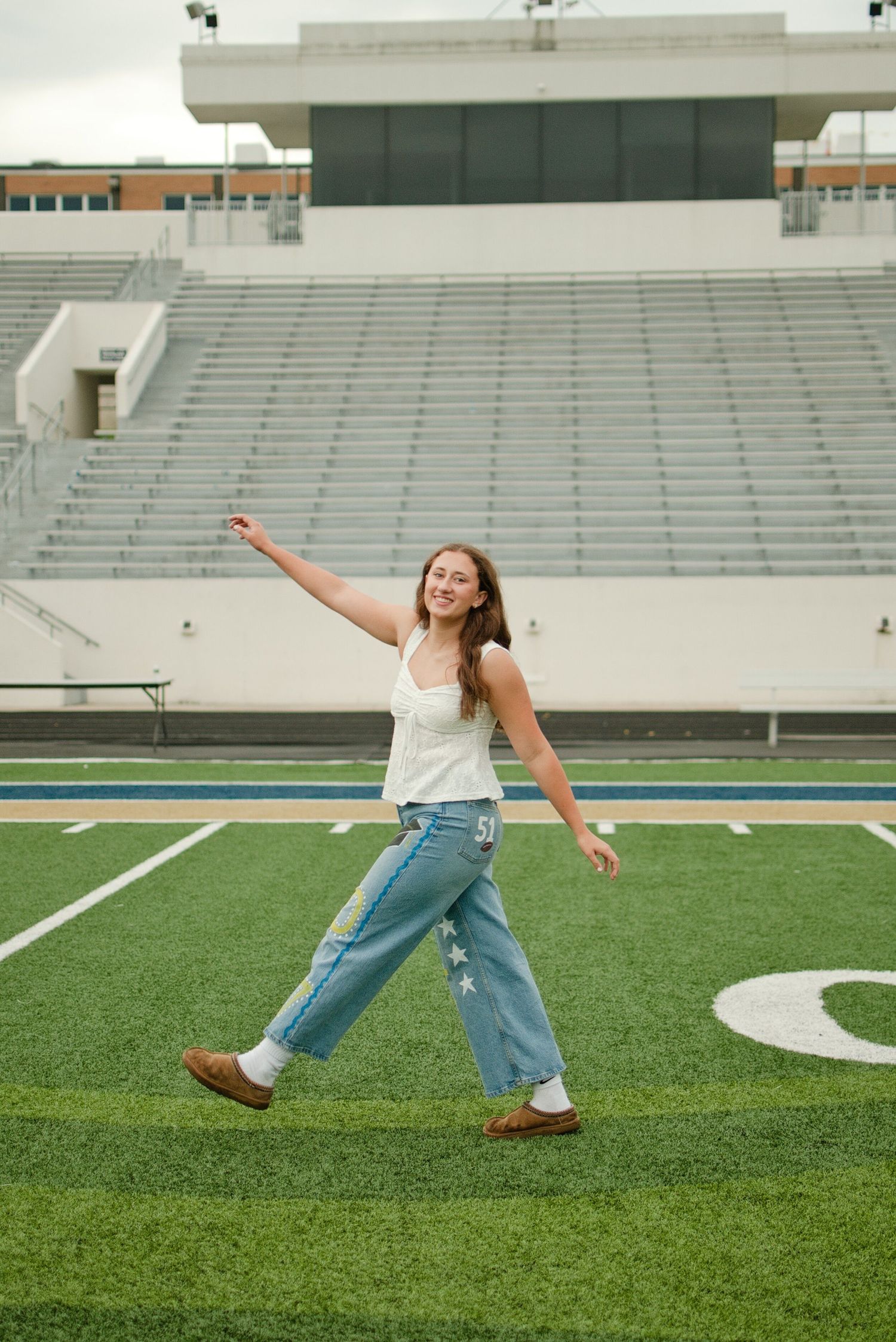 Young woman in white top and jeans walks on football field with stadium bleachers in background.