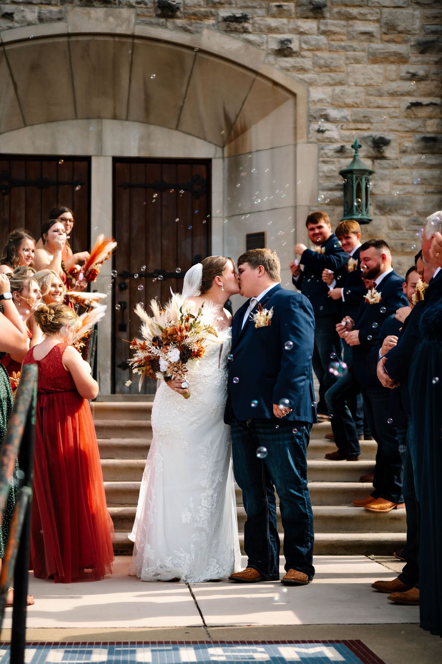 Wedding party celebrates outside stone church entrance with bridesmaids in coral dresses.