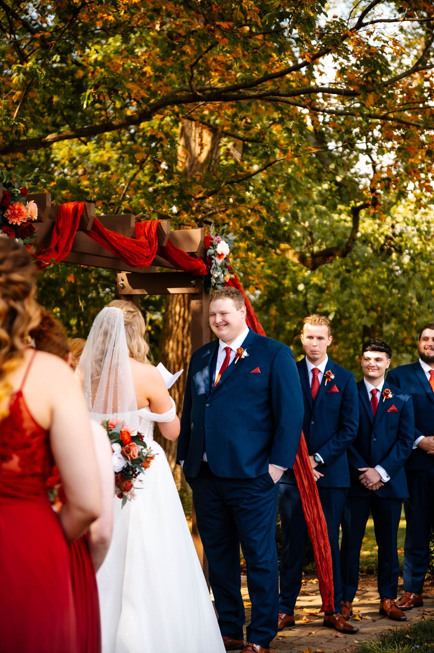 Wedding party in navy suits and red dresses pose during outdoor autumn ceremony.
