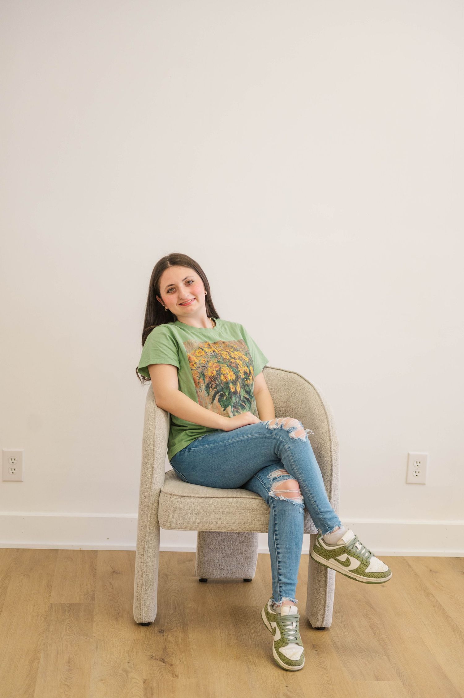 Smiling brunette in green graphic tee and ripped jeans seated in modern chair with green sneakers.