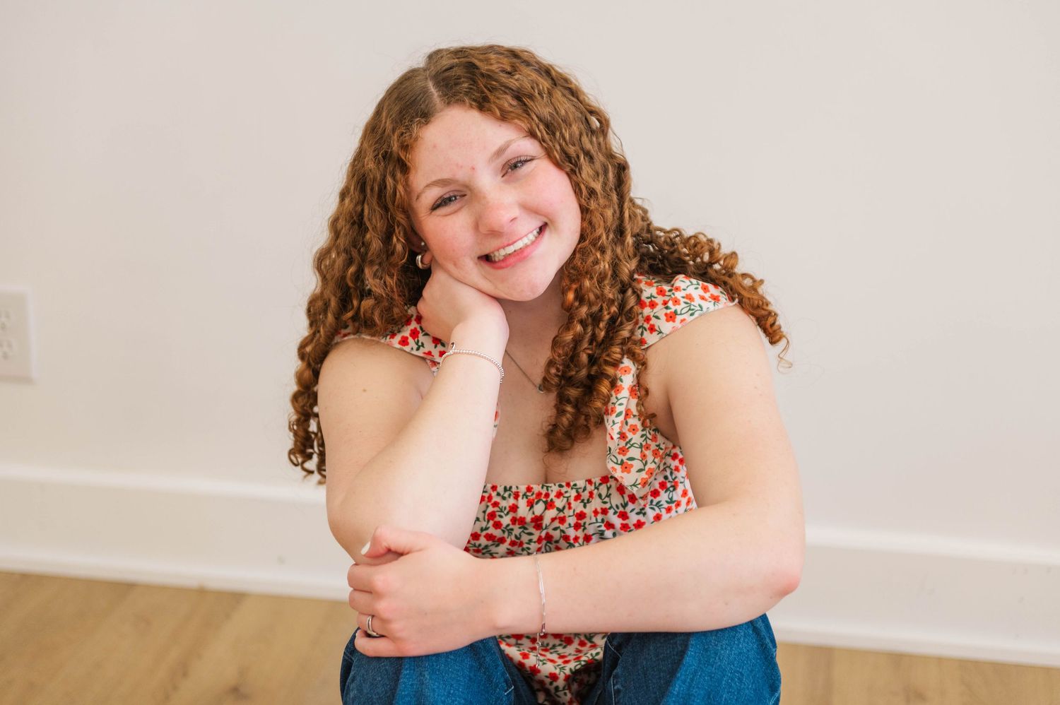 Young girl with curly brown hair smiling while sitting on floor in colorful floral top.
