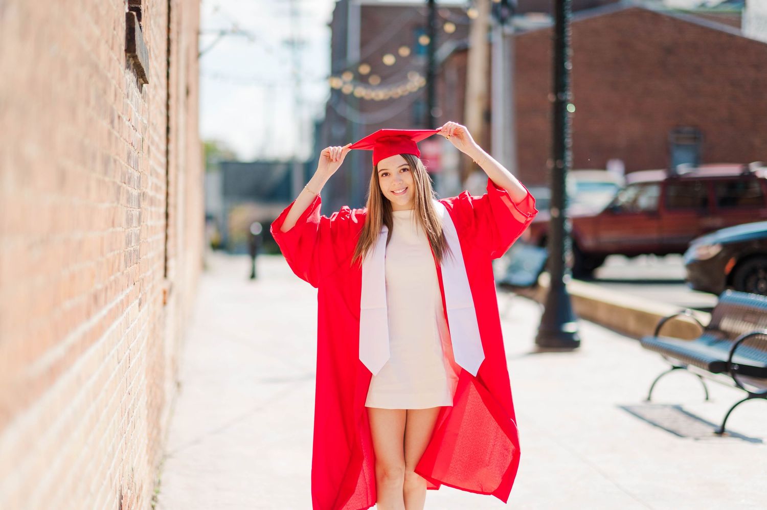 A smiling graduate poses outdoors in a red cap and gown on a city sidewalk on graduation day.