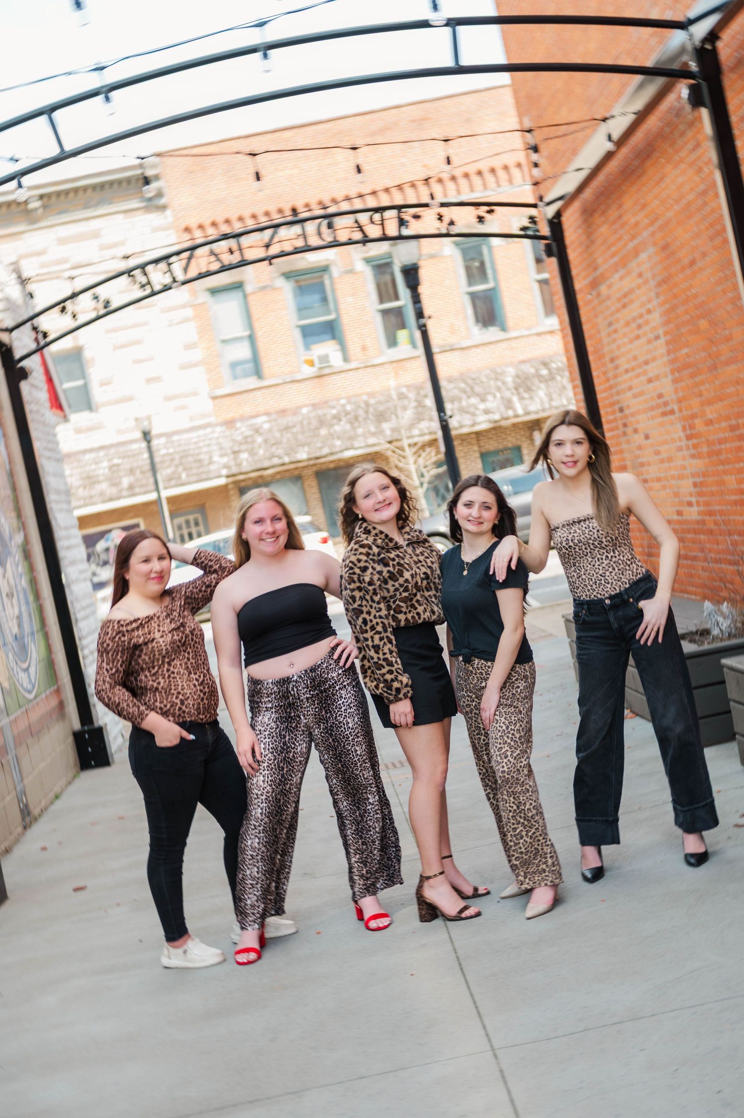 Five smiling young women in leopard print and black outfits posing together under an arch on a city sidewalk.
