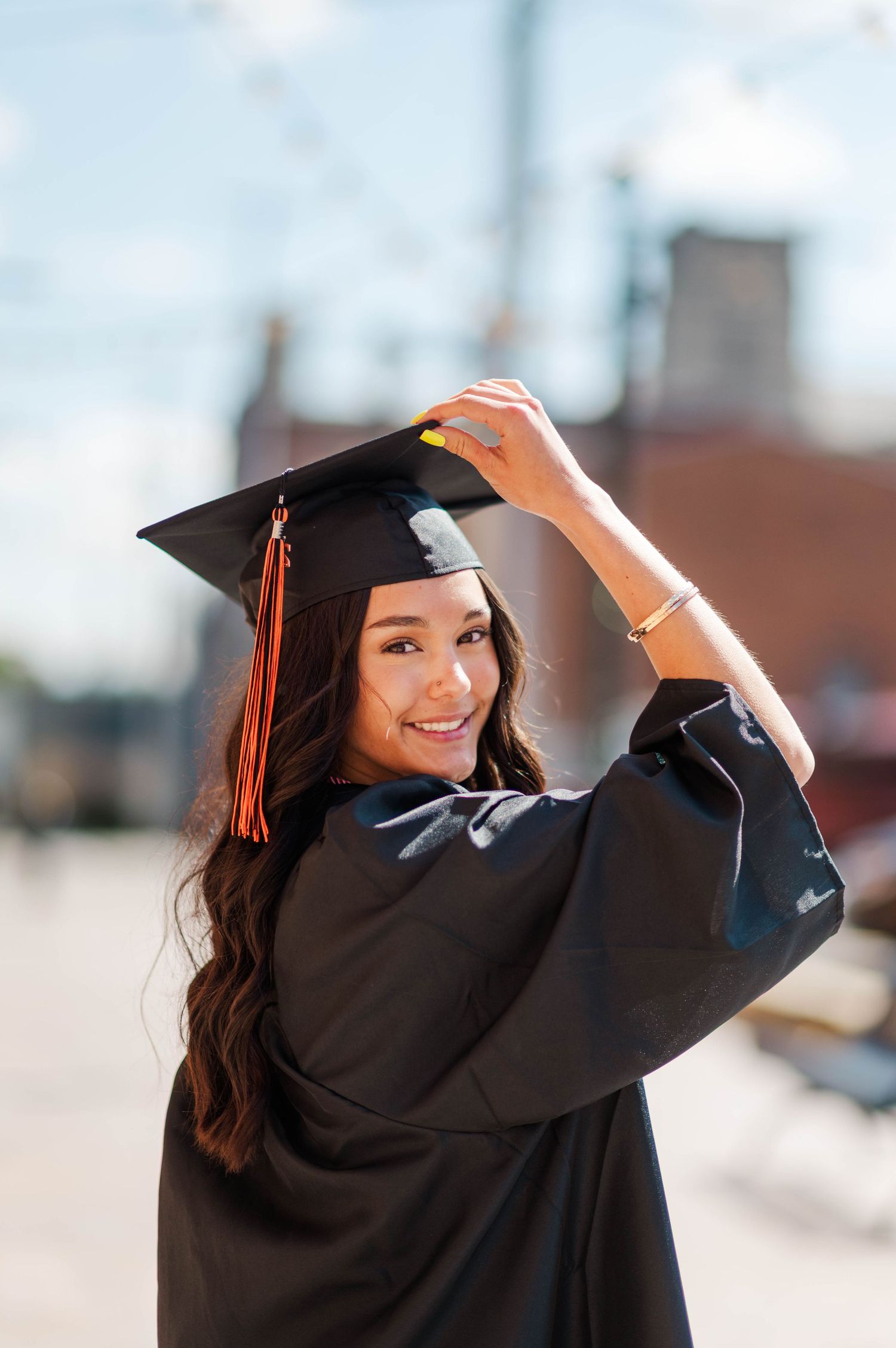 Smiling graduate in black cap and gown adjusts her mortarboard against a bright blue sky.