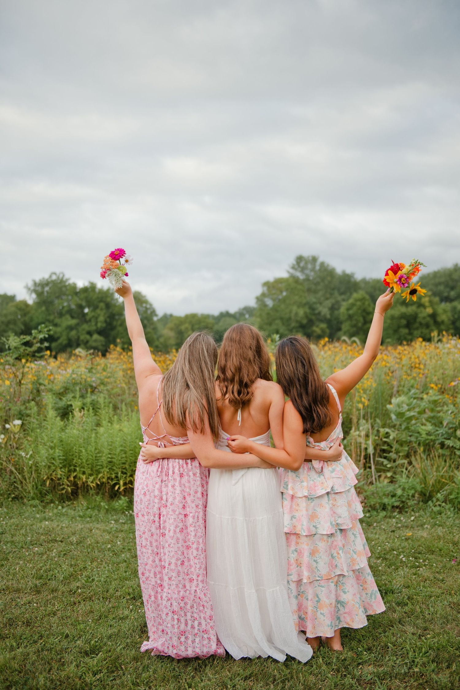 Three women in floral dresses embrace outdoors, raising bouquets of colorful flowers in a sunny garden setting.