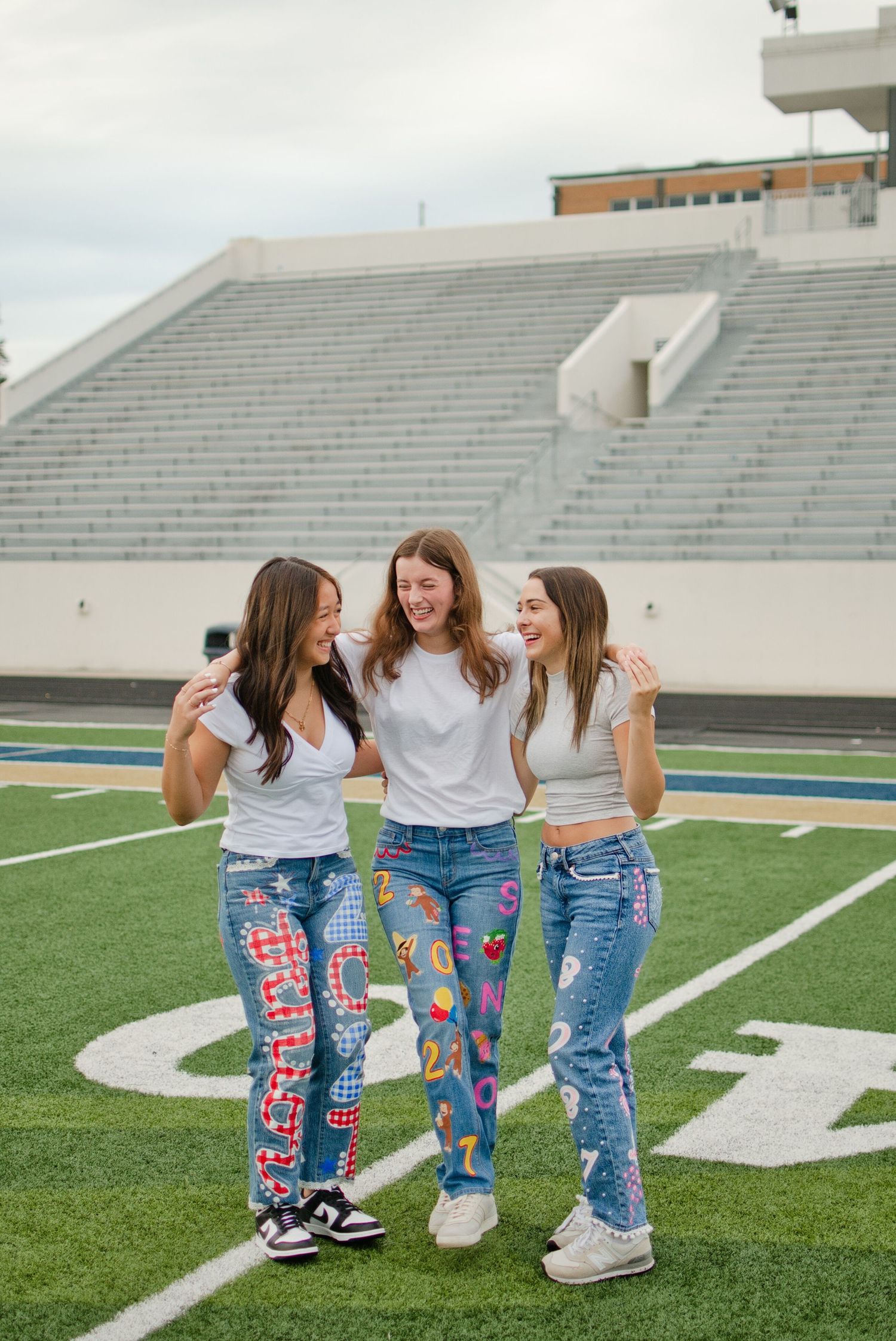 Three smiling teen girls in colorful custom painted jeans laugh together on a green football field.
