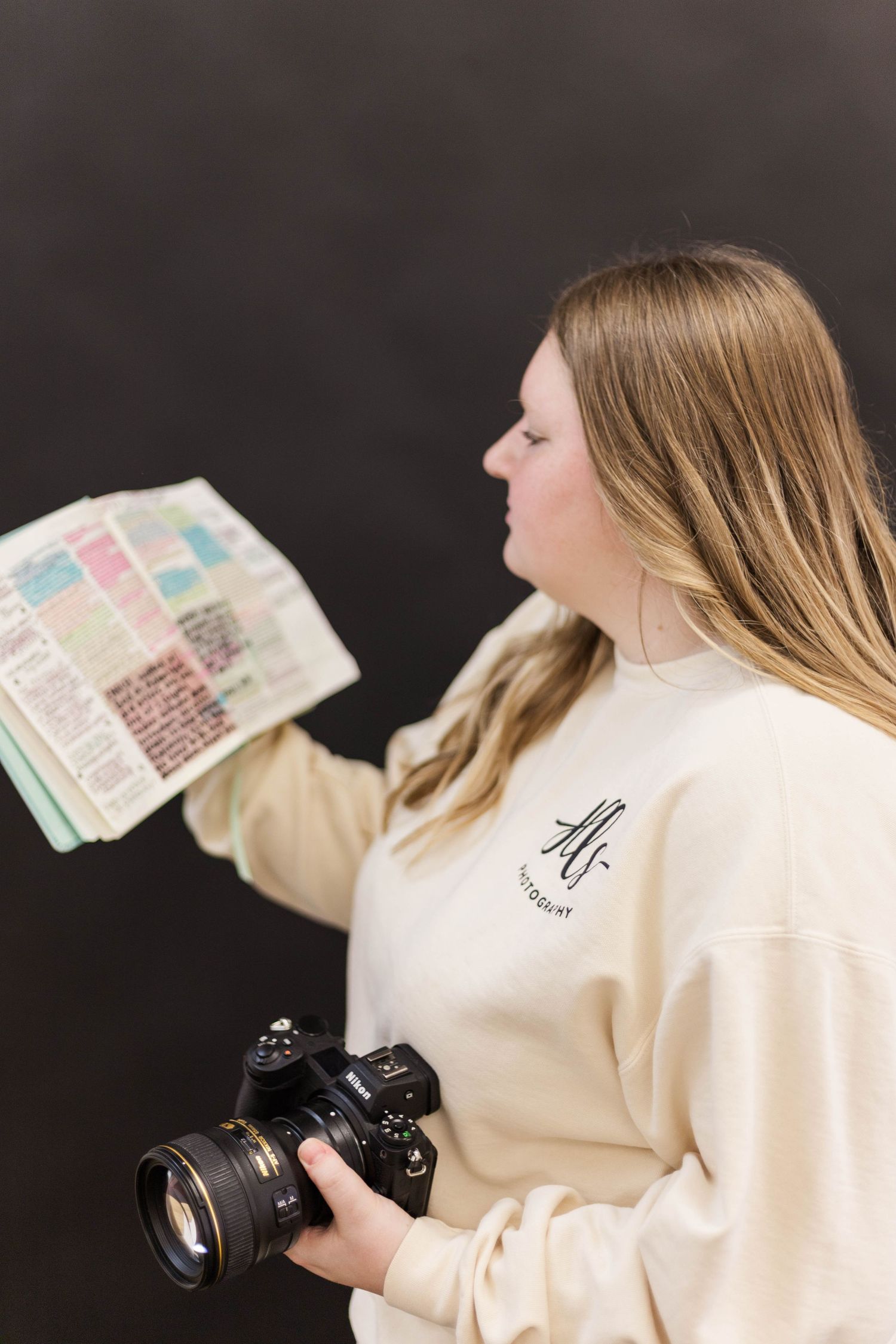 Photographer in cream sweater holds Bible and camera against dark background.
