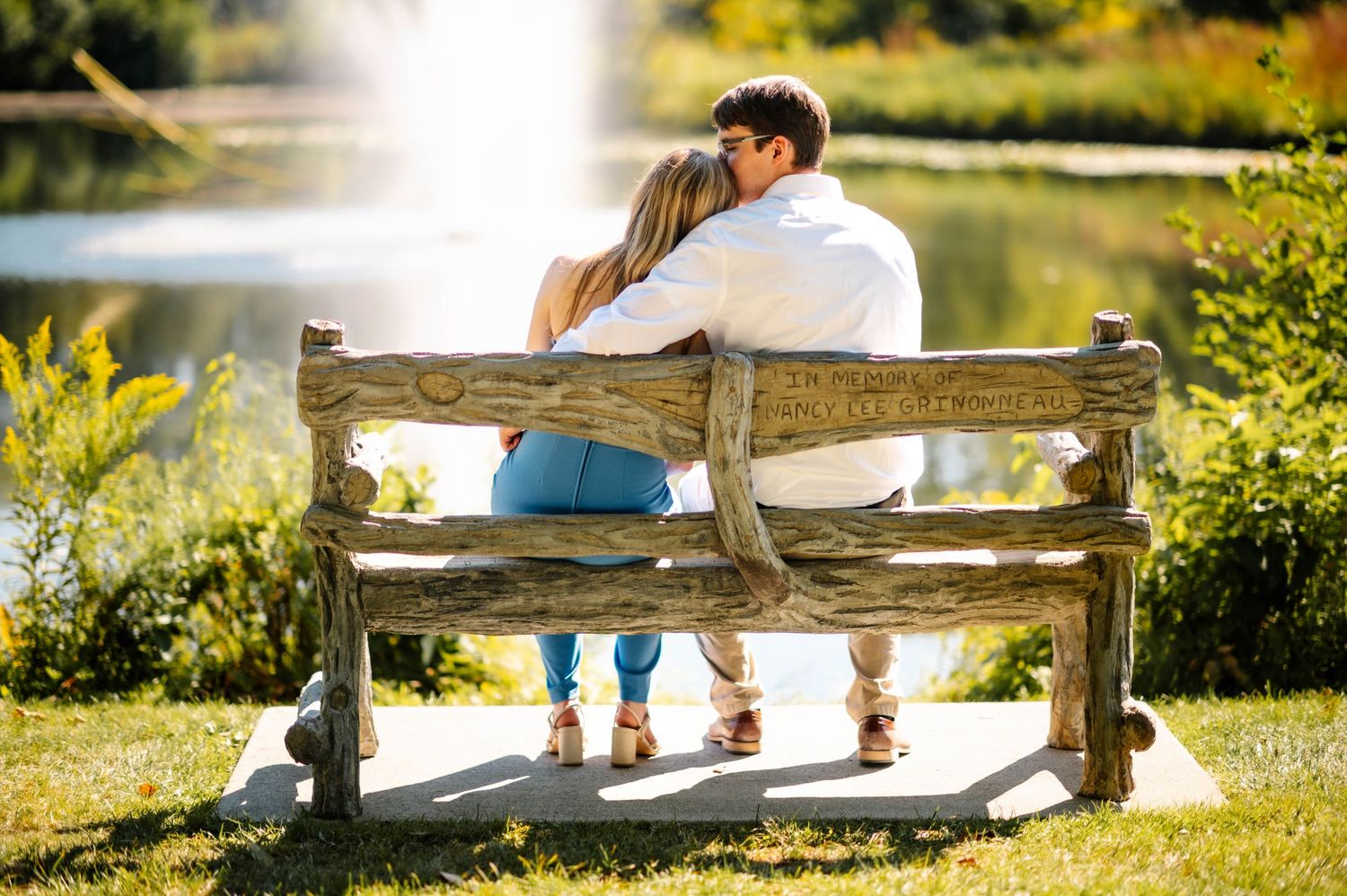 A couple sitting close together on a wooden bench overlooking a serene pond with fountain.