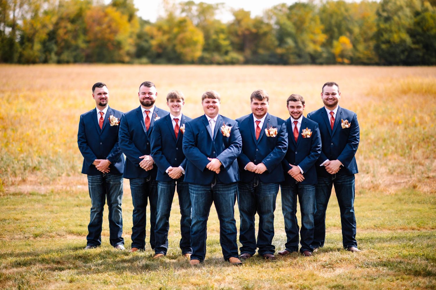 Wedding party members in navy suits pose playfully in a golden autumn field at sunset.