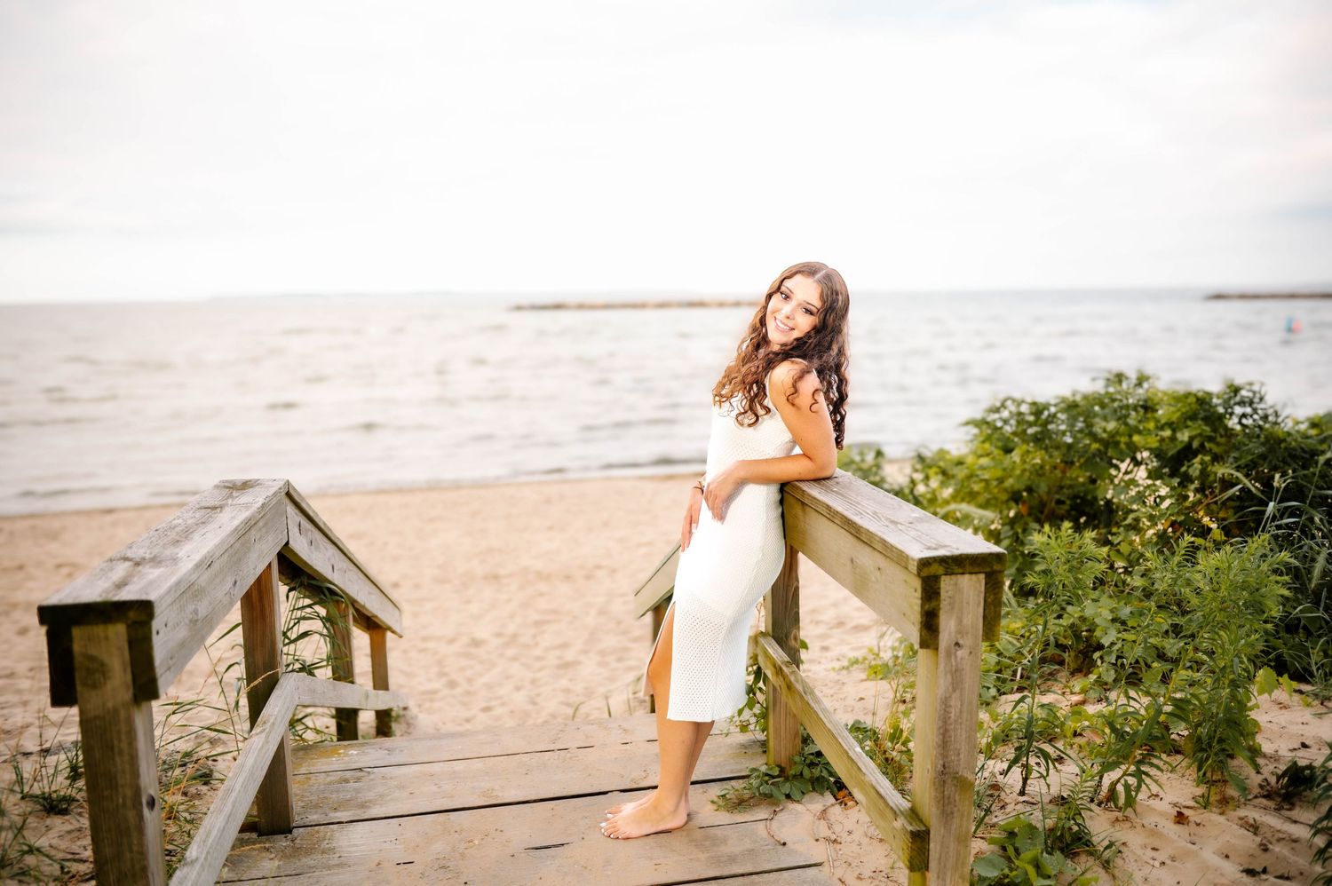 A young woman in a white dress poses on wooden beach boardwalk steps with sandy shore and calm sea behind her.