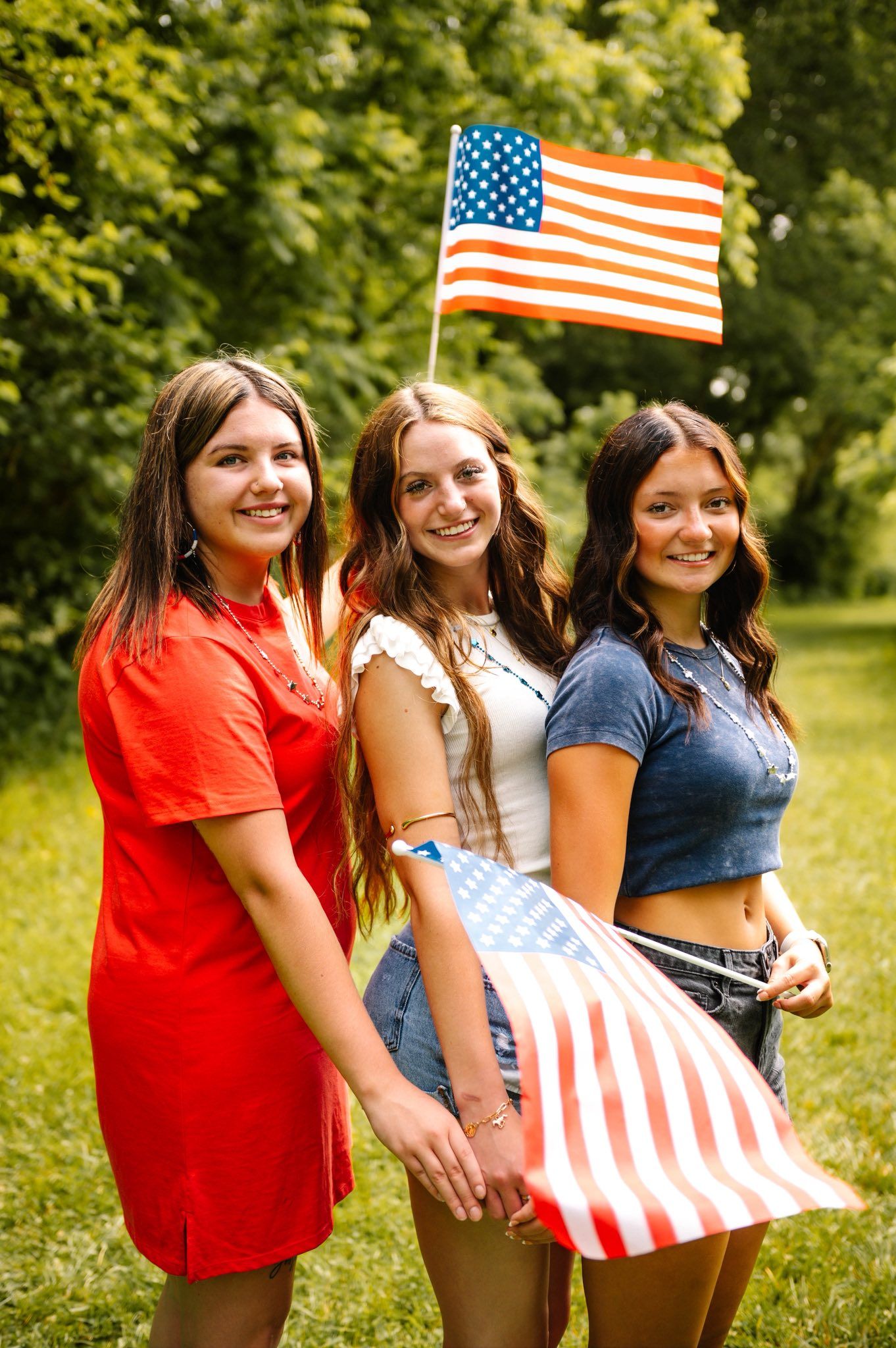 Three friends in patriotic outfits pose with American flags in sunny outdoor setting.