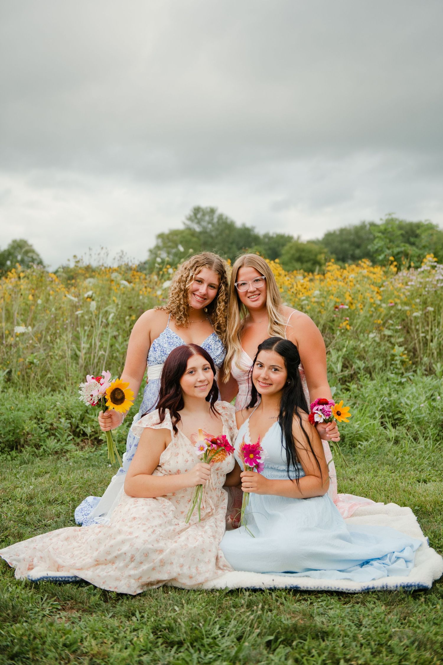 Four women in floral dresses pose together in a wildflower field holding colorful bouquets on a cloudy day.