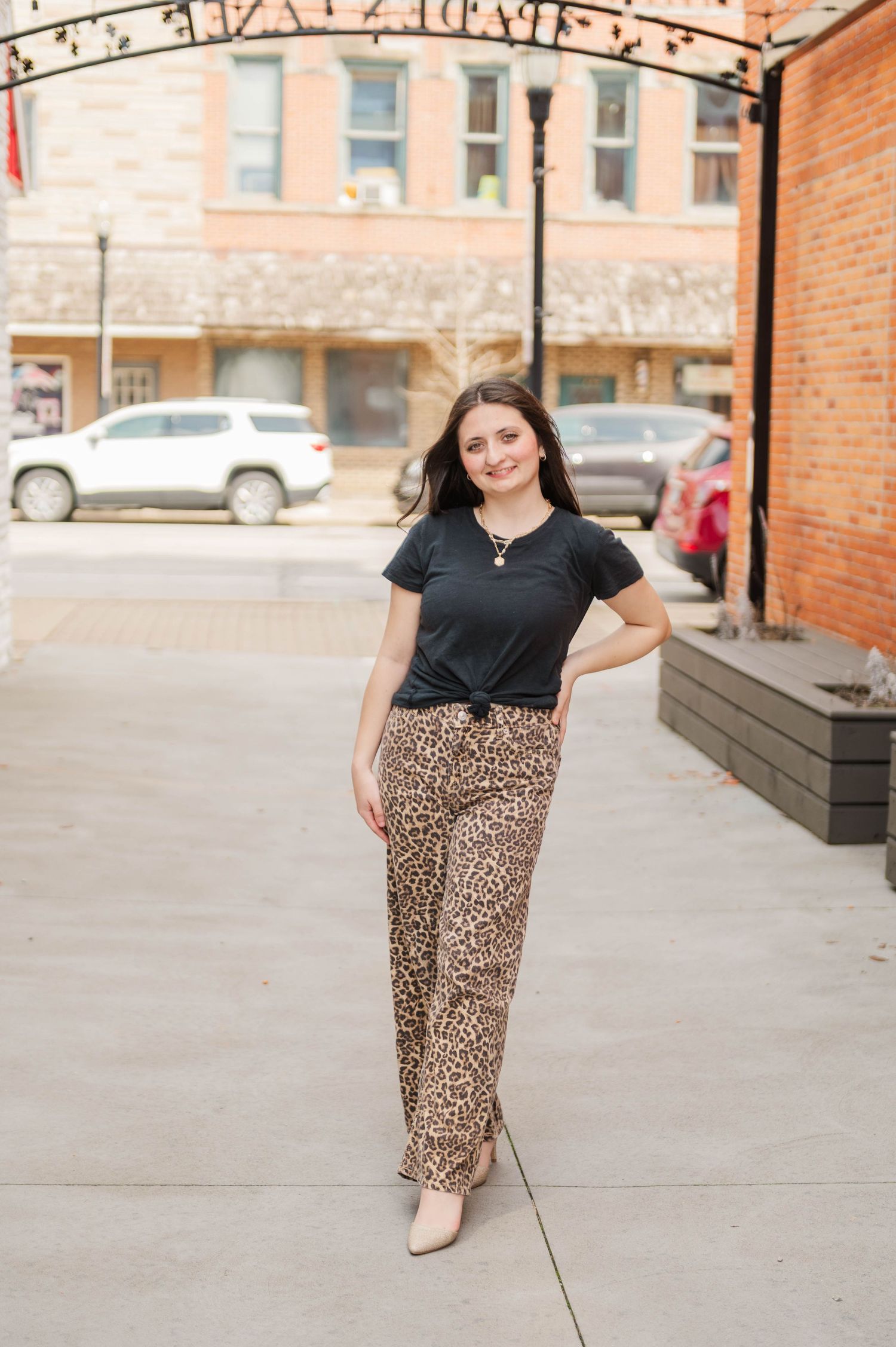 Young woman in black t-shirt and leopard print pants poses confidently under decorative arch outdoors.