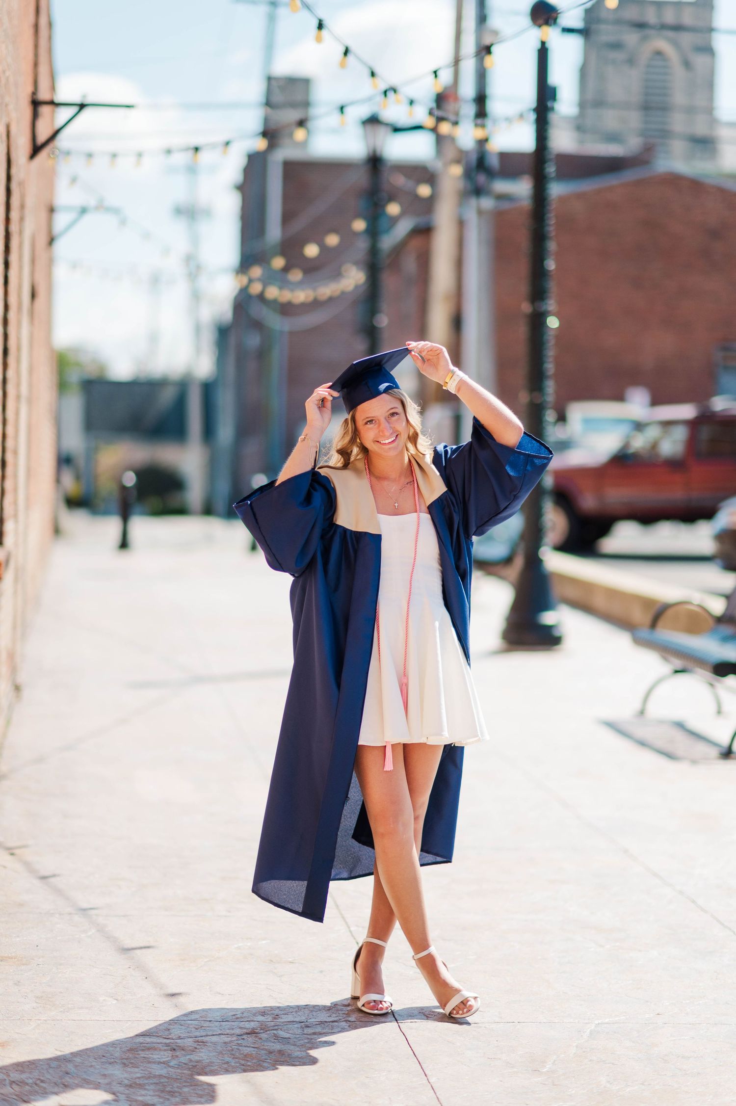 Smiling graduate in navy cap and gown poses on a sunny sidewalk with string lights above.