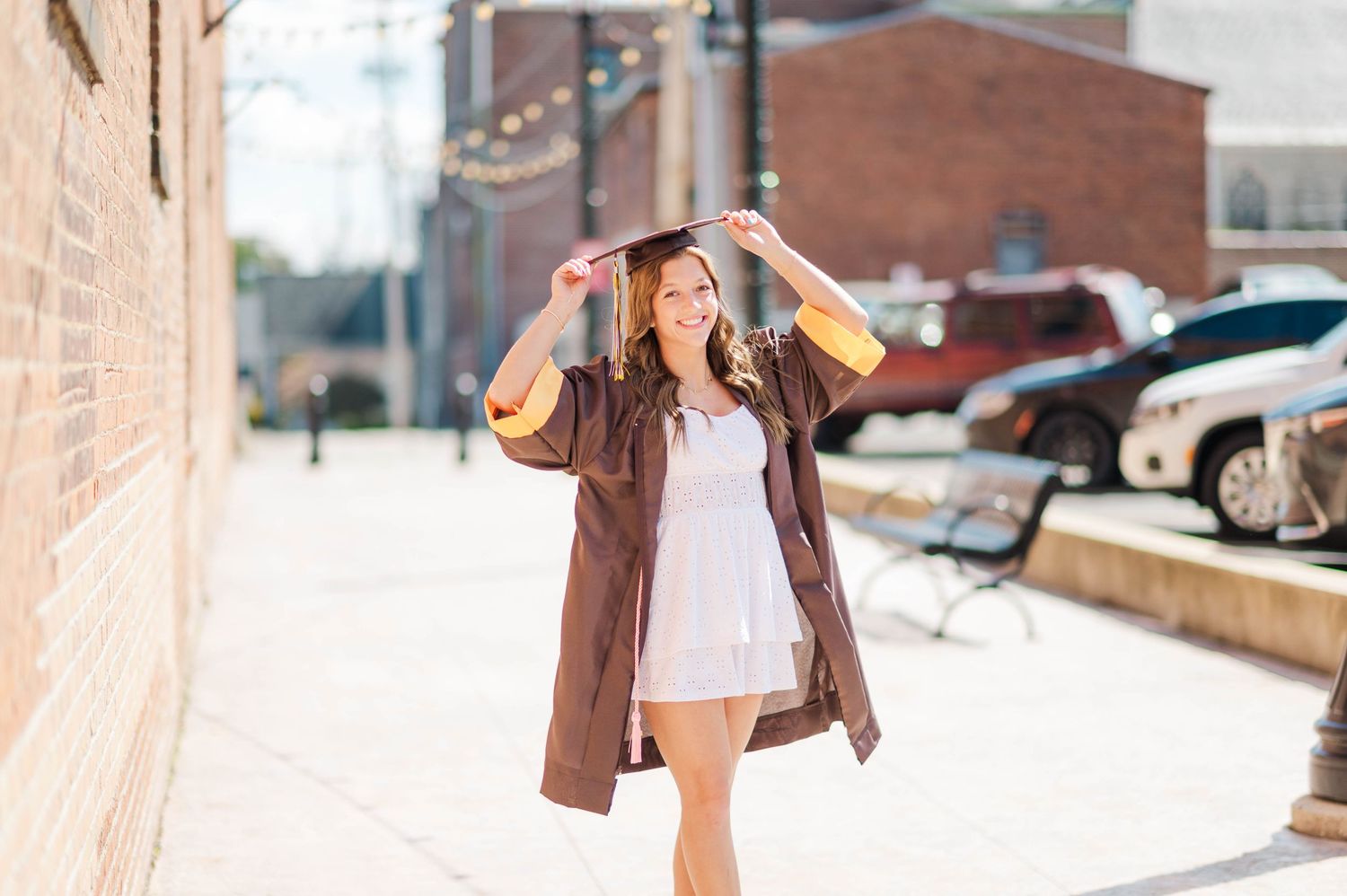 Happy graduate in brown and gold cap and gown walking outdoors adjusting her cap on sunny day.