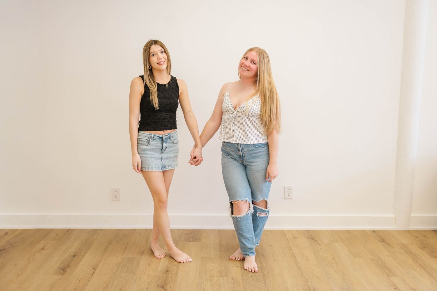 Two women in casual outfits hold hands while standing barefoot against a white wall indoors.