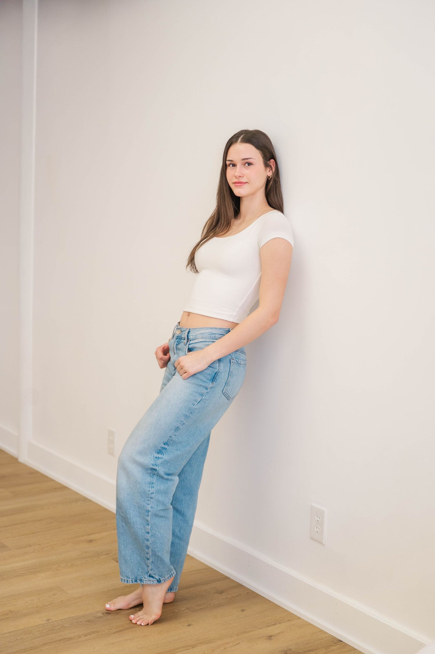 Brunette woman in white crop top and blue jeans standing barefoot against white wall on hardwood floor.