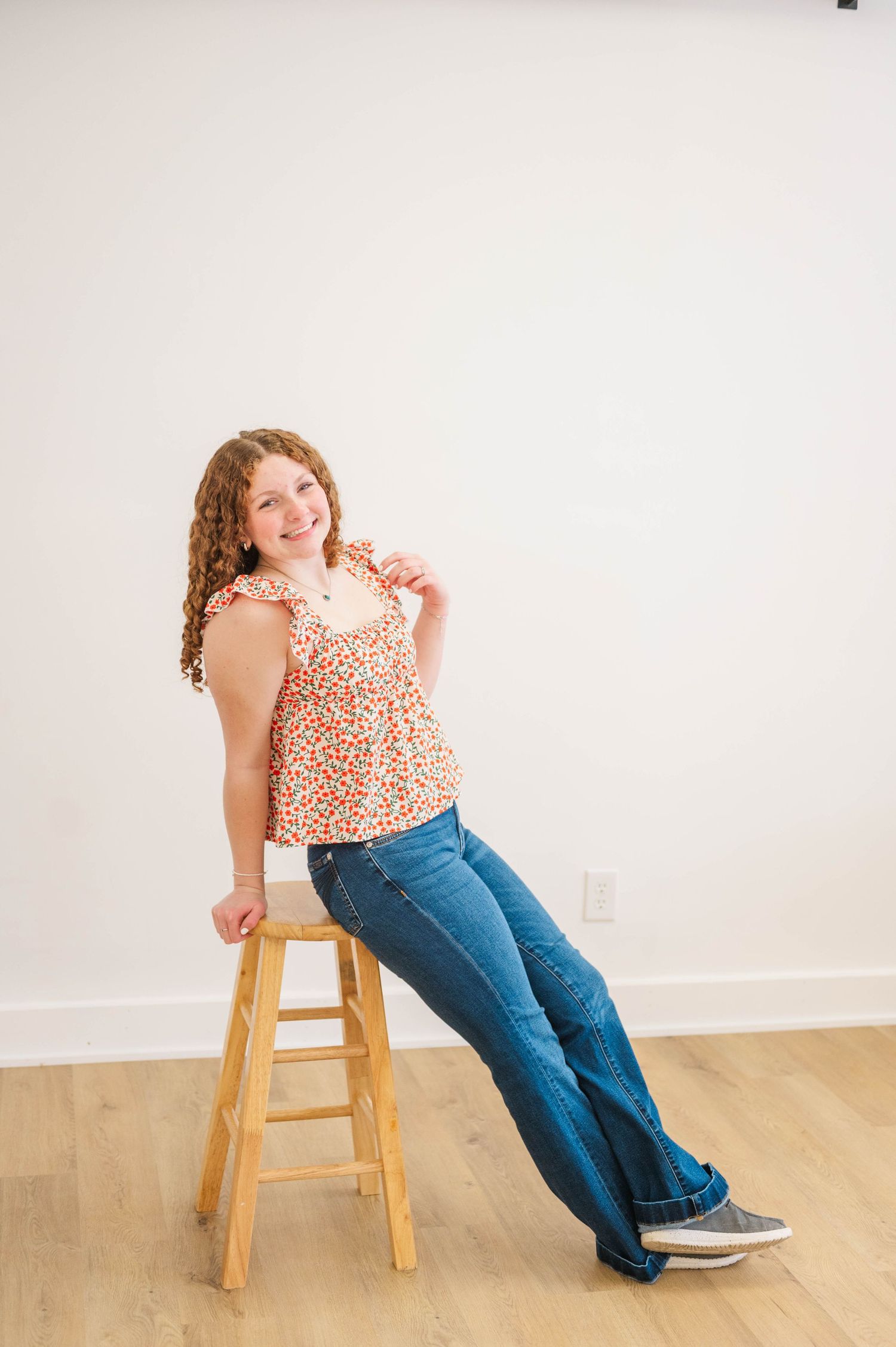 Young girl with curly hair sits on wooden stool wearing floral top and flare jeans, smiling at camera.