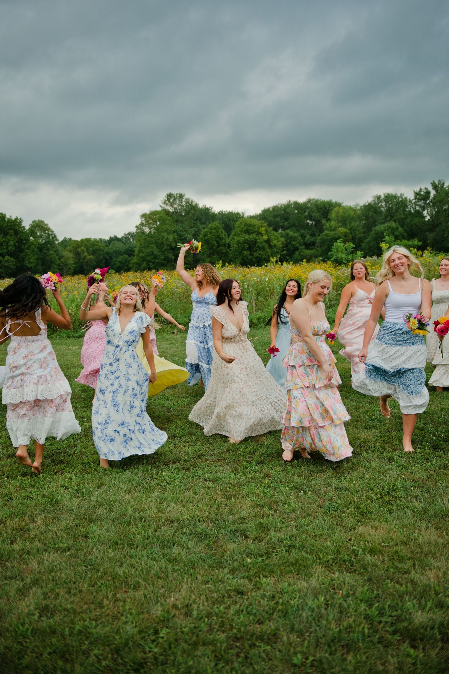 Joyful bridesmaids in floral dresses running through a green field on a cloudy day.