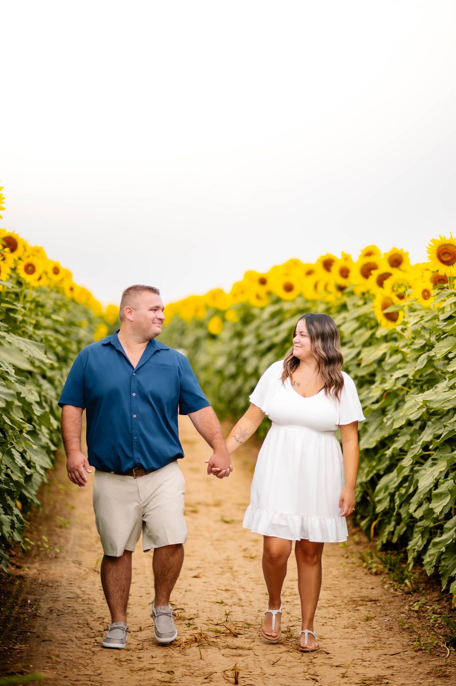 A couple walking hand in hand through rows of tall sunflowers on a dirt path.
