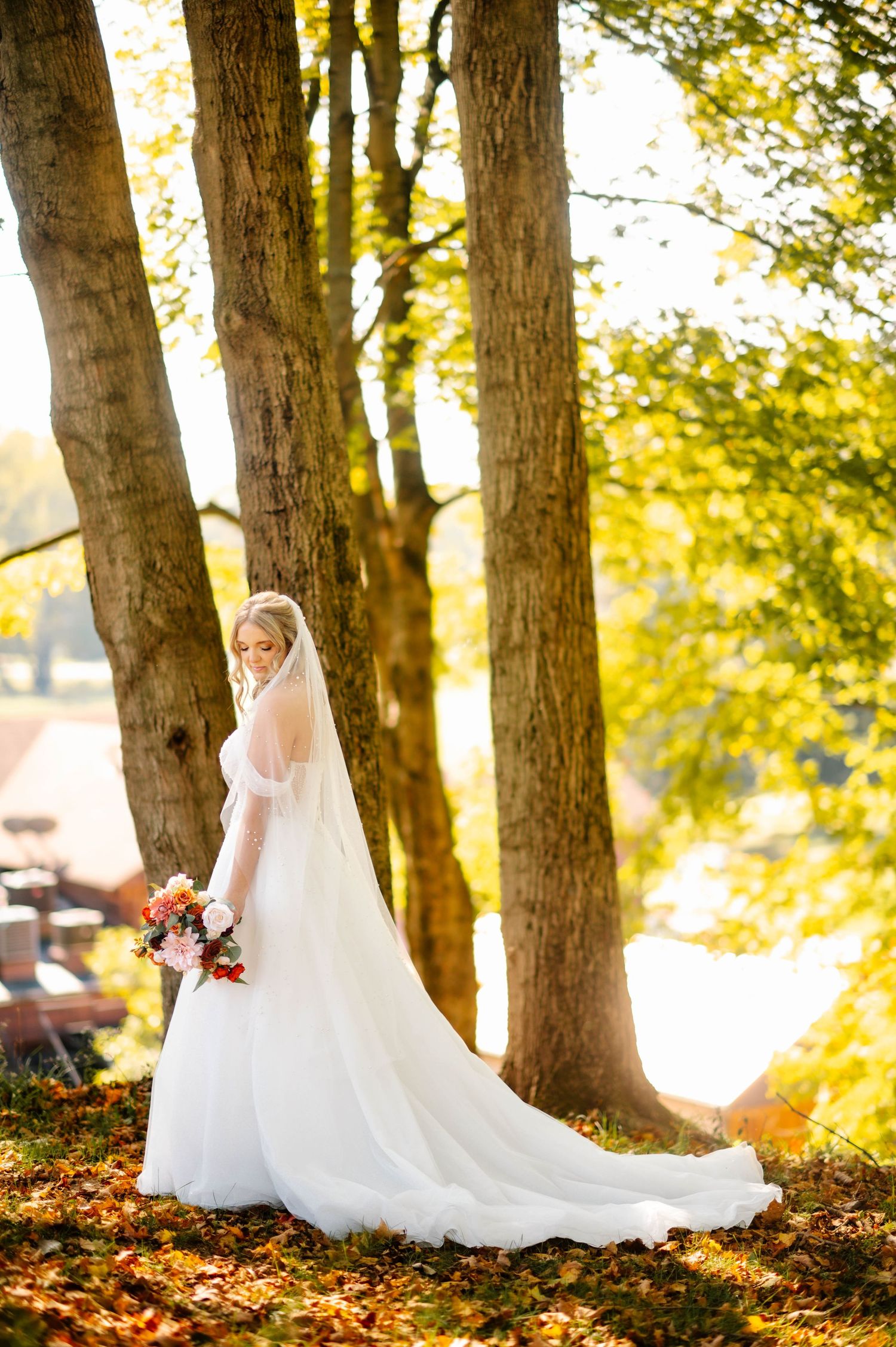 Bride in flowing white gown poses among autumn trees with golden sunlight filtering through.