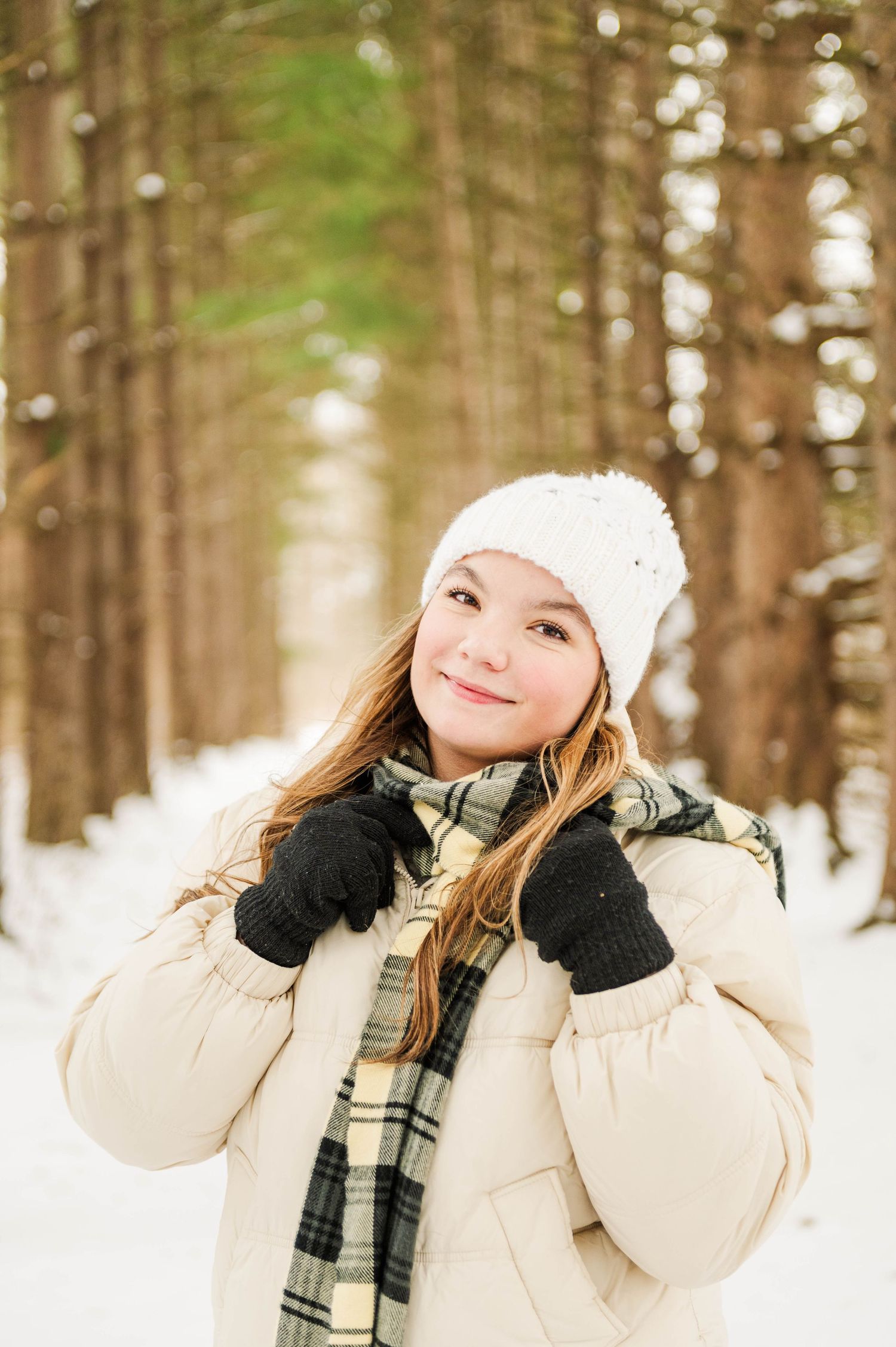 Person in cream puffer jacket and white beanie poses in snowy forest setting.