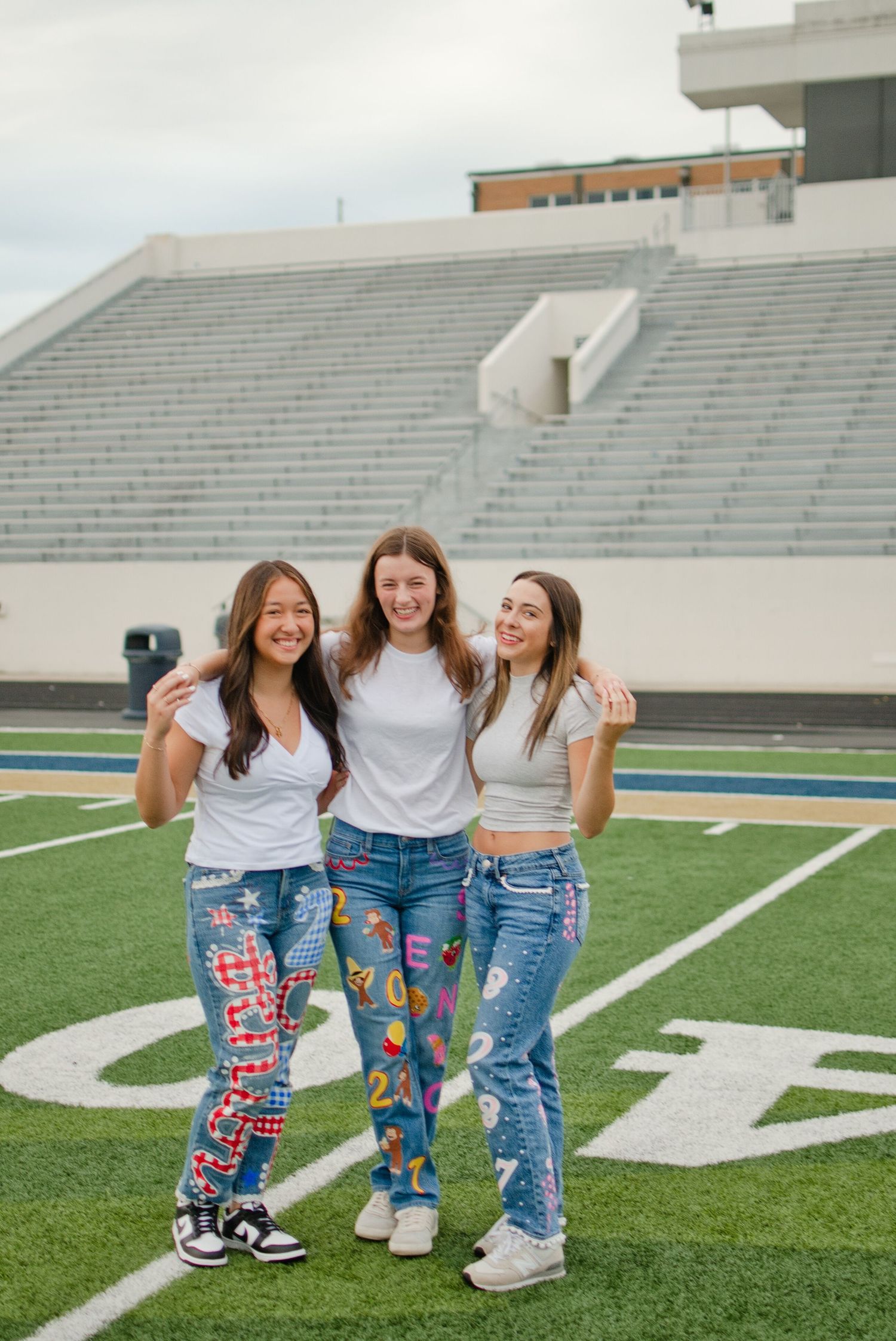 Three teen girls in custom painted jeans and white tees pose together smiling on a football field.