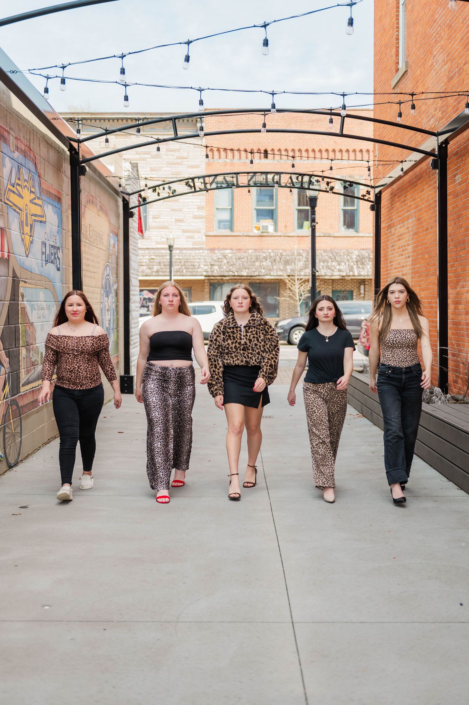 Five young women in animal print outfits walk confidently toward camera under string lights in urban alley.