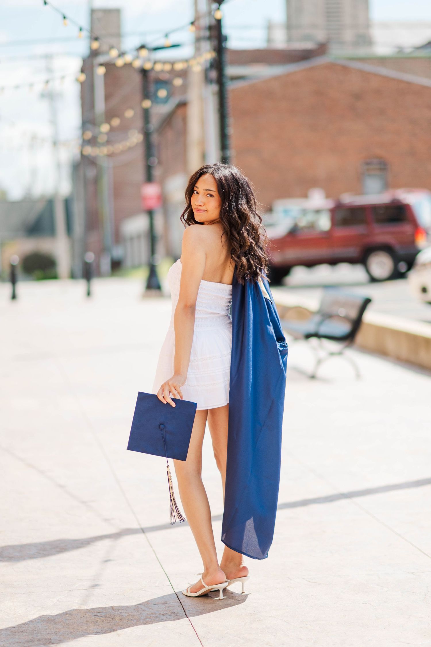 Brunette woman in white dress holding navy graduation cap and gown on sunny urban street.
